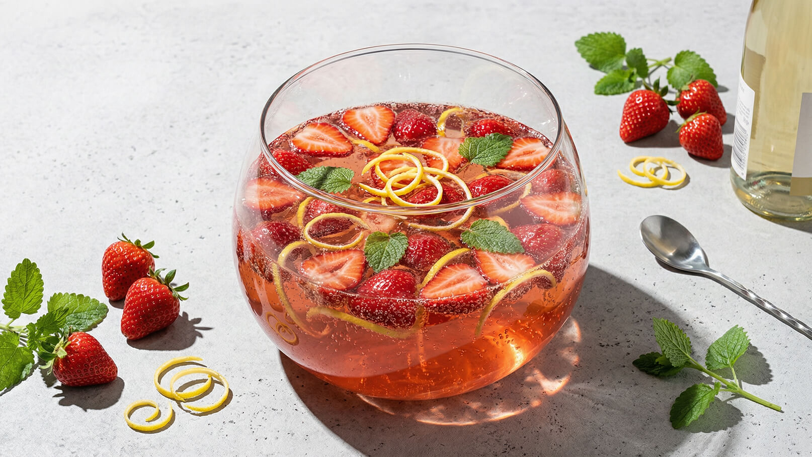 Eine große Glasbowle enthält sprudelndes Getränk mit Erdbeeren, Zitronenschalen und frischen Minzblättern. Um die Bowle herum liegen weitere Erdbeeren und Minzblätter auf einer hellen Oberfläche, während ein Löffel und einige Zitronenschalen daneben platziert sind.