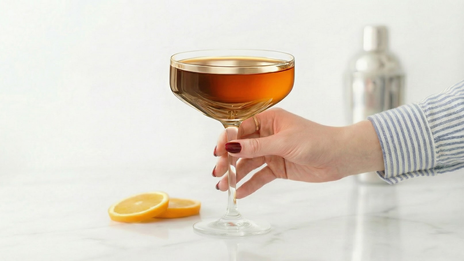 Eine Hand hält ein elegantes Cocktailglas mit einer goldbraunen Flüssigkeit. Neben dem Glas liegen zwei Scheiben Zitrone auf einer hellen Oberfläche. Im Hintergrund ist ein Shaker zu erkennen.