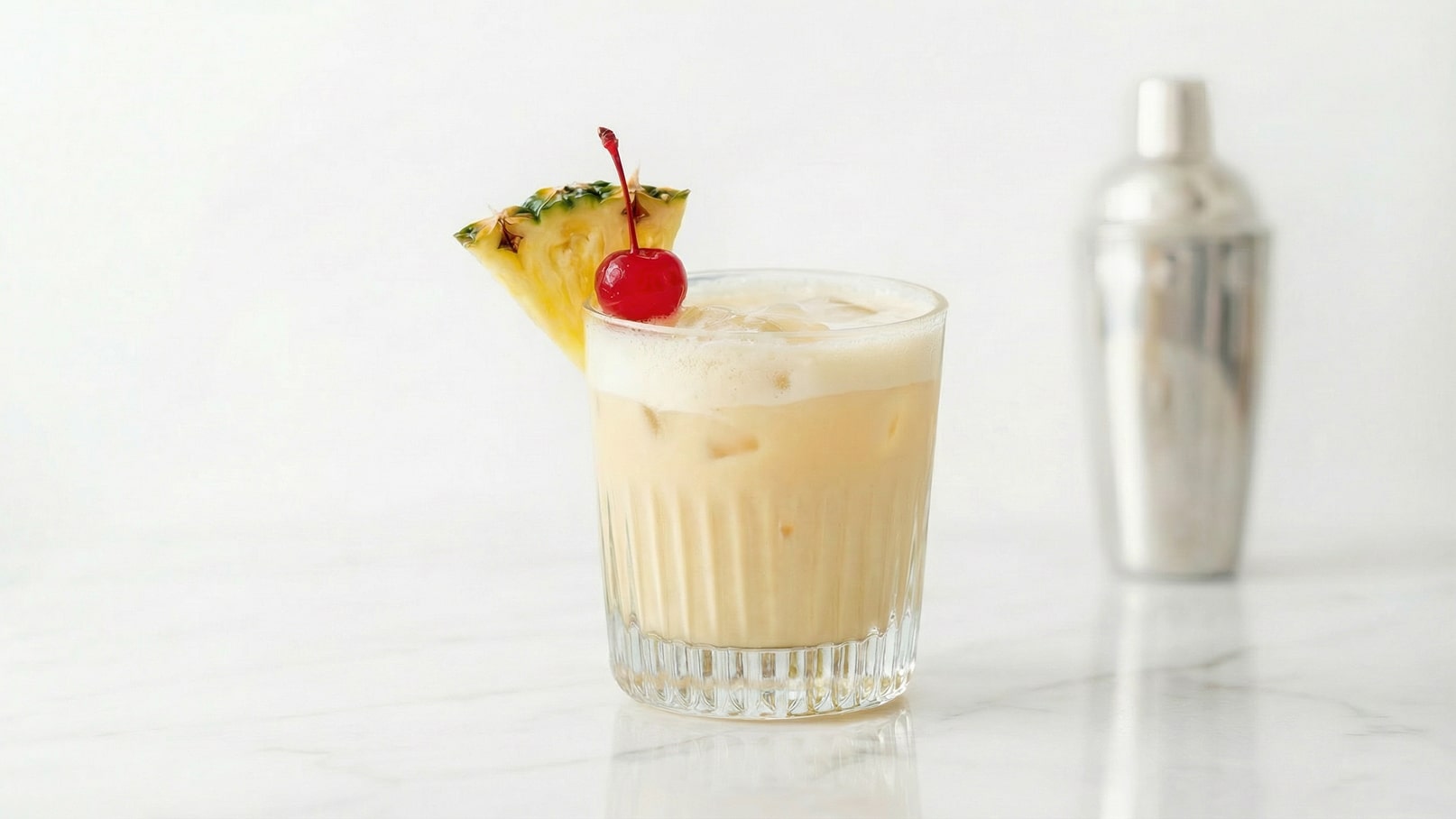 Ein Cocktail in einem klaren Glas mit Eiswürfeln, garniert mit einer Ananasscheibe und einer roten Kirsche. Im Hintergrund steht ein silberner Shaker. Der Cocktail hat eine cremige, hellbeige Farbe.