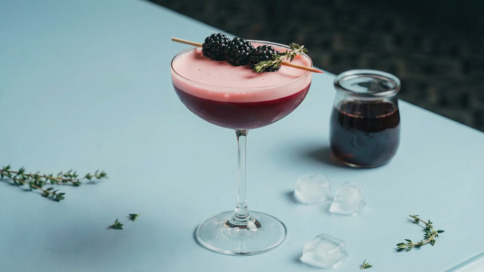 Ein eleganter Cocktail in einem hohen Glas mit einem rosa Schaum und dunkler Flüssigkeit darunter. Oben sind frische Brombeeren und ein Zweig Thymian dekoriert. Daneben steht ein kleines Glas mit einer dunklen Flüssigkeit und einige Eiswürfel auf dem Tisch.