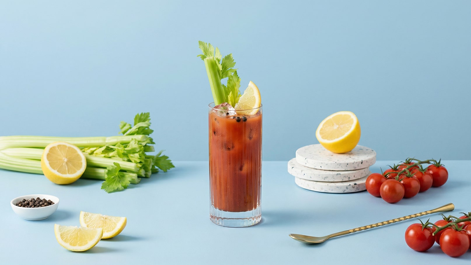Ein hoher Glas mit einem roten Getränk, garniert mit Sellerie und Zitronenscheibe. Daneben liegen frische Zitronen, eine Schale mit Pfefferkörnern, eine kleine Ansammlung von Kirschtomaten und einige Selleriestangen auf einem blauen Hintergrund.
