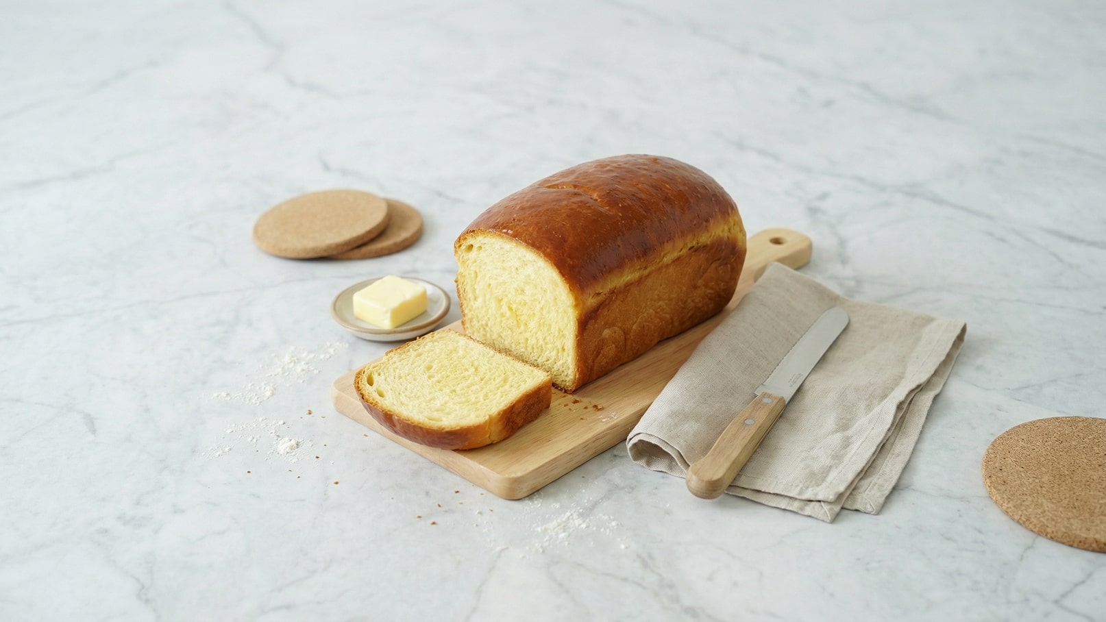 Ein frisch gebackenes, goldbraunes Brot steht auf einem Holzbrett. Es ist teilweise aufgeschnitten, und neben dem Brot liegt ein Butterstück auf einem kleinen Teller. Ein Messer aus Edelstahl liegt auf einer stofflichen Unterlage. Der Hintergrund ist hell und neutral.