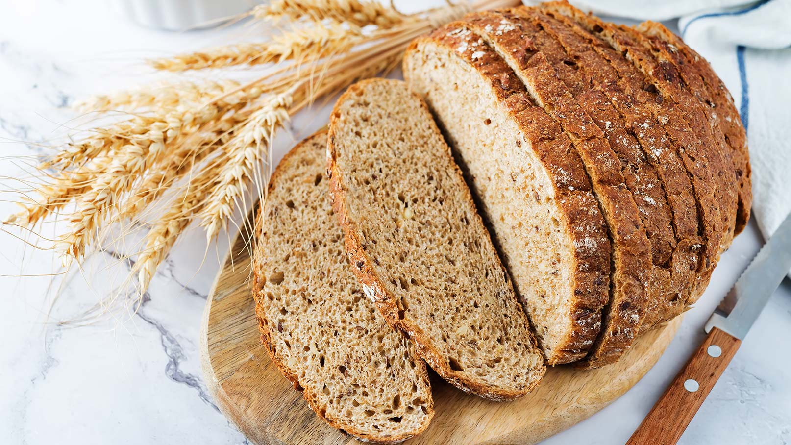 Auf einem rustikalen Holzbrett liegt ein geschnittener Laib Roggen-Kartoffelbrot, umgeben von getrockneten Getreidekörnern. Ein traditionelles Messer liegt neben dem Brot, das auf einem marmorierten Stein ruht. Das Bild strahlt Natürlichkeit und Handwerkskunst aus.