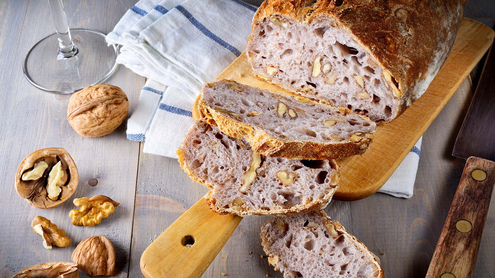 Auf dem Bild ist ein saftiges Walnussbrot auf einem rustikalen Holzbrett zu sehen. Der vordere Teil des Brotes ist bereits angeschnitten, und die saftige Krume und die knackigen Walnussstücke sind deutlich zu erkennen. Zur Dekoration des Brotes liegen einige ganze Walnüsse neben dem Brot. Unter dem Holzbrett liegt ein weiß-blau kariertes Tuch, das dem Ganzen eine heimelige und gemütliche Note verleiht.