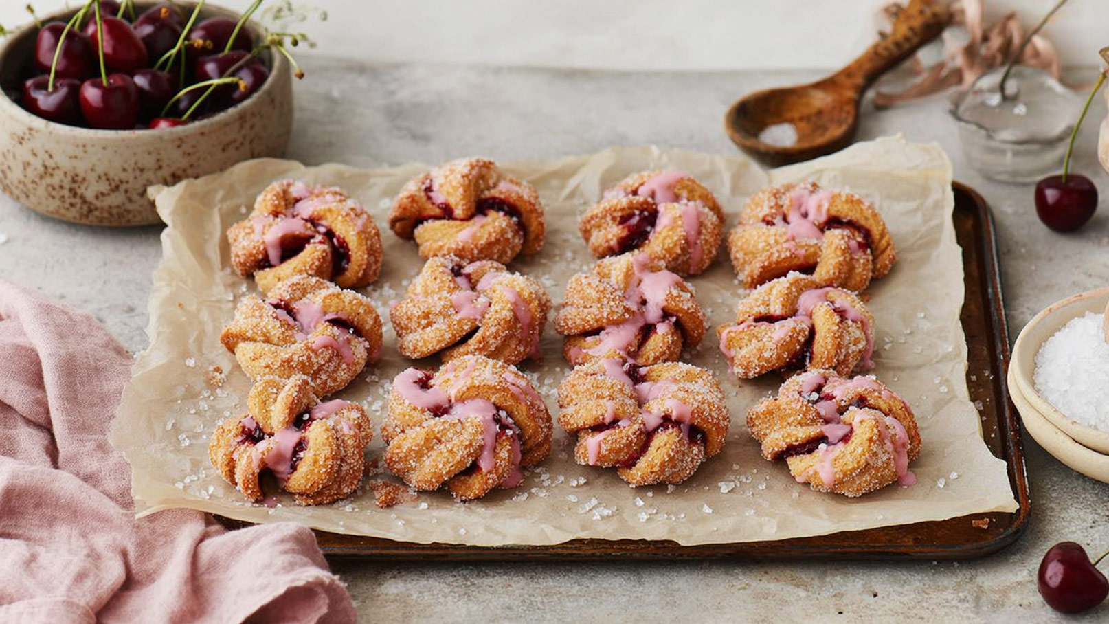 Eine Auswahl von gebackenen Süßigkeiten mit einer rosa Glasur und Zucker bestreut, die auf einem Backblech liegen. Im Hintergrund sind Kirschen und eine kleine Schüssel mit Salz zu sehen. Die Leckereien haben eine knusprige Textur und eine fruchtige Füllung.