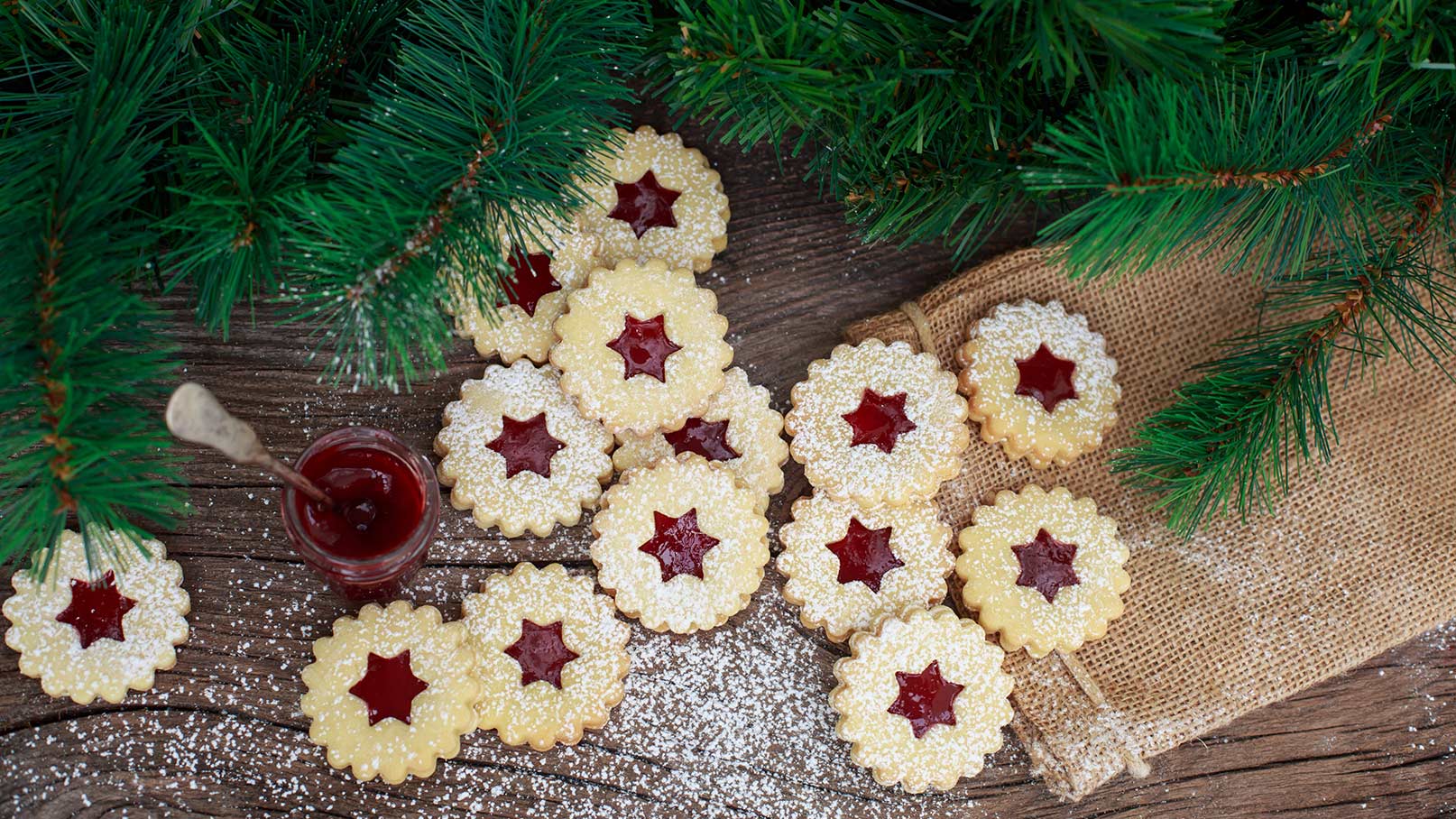 Das Bild zeigt einen rustikalen Holztisch, der mit zahlreichen Linzer Augen dekoriert ist. Diese traditionellen Kekse sind liebevoll angerichtet und glänzen mit einer feinen Schicht Puderzucker, die ihnen eine süße Note verleiht. Im Hintergrund ragen grüne Tannenzweige hervor, die eine festliche, winterliche Atmosphäre schaffen.