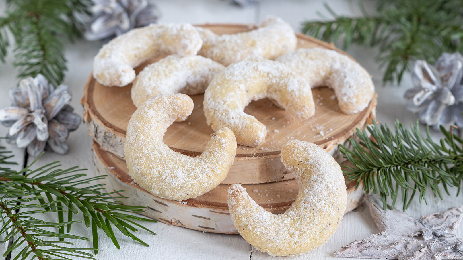 Das Bild zeigt rustikale Holzplatten mit köstlichen Vanillekipferln vom Donau-Kurier, die mit Puderzucker bestäubt sind. Neben den Platten liegt ein grüner Tannenzweig, der eine festliche, weihnachtliche Atmosphäre schafft. Die Präsentation strahlt Gemütlichkeit und Genuss aus.
