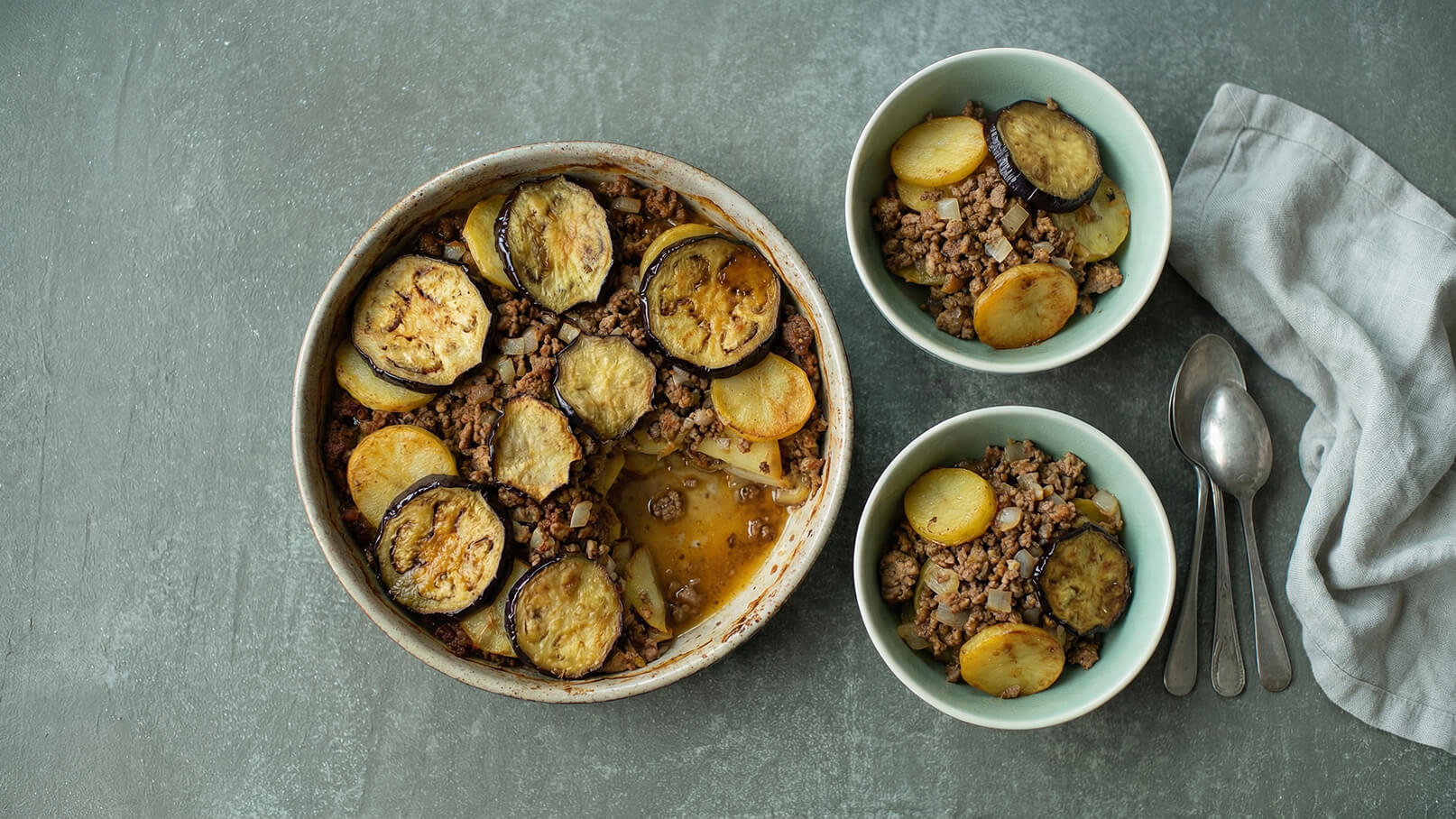 Ein tiefes Gericht mit gebackenem Hackfleisch, Auberginen und Kartoffelscheiben ist in der Mitte zu sehen. Daneben stehen zwei kleine Schalen, die ebenfalls mit Hackfleisch, Auberginen und Kartoffeln gefüllt sind. Ein paar Löffel liegen daneben.