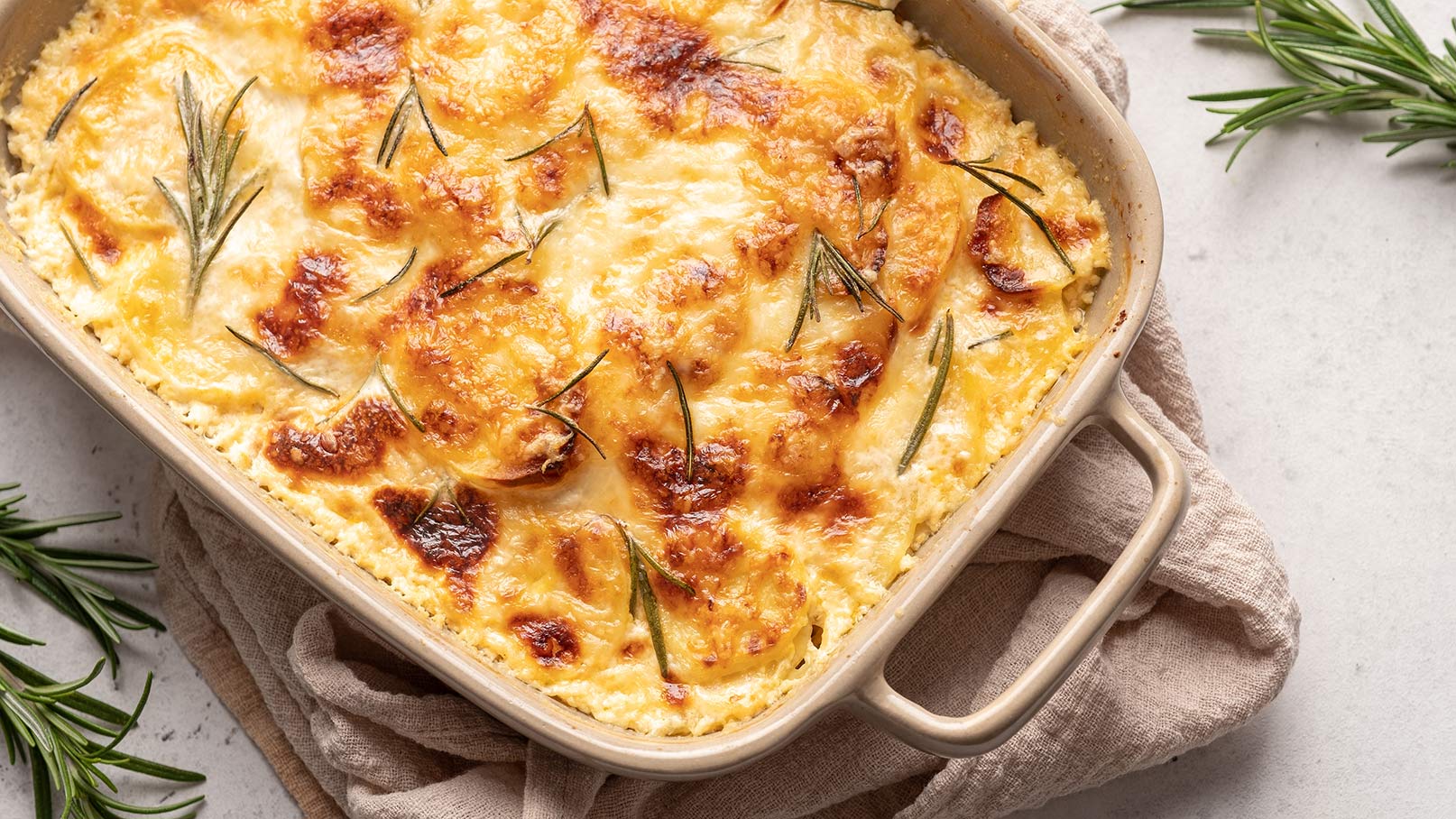 Das Bild zeigt einen köstlichen Kartoffel-Käse-Auflauf, der mit Rosmarin bestreut ist. Der Auflauf ist schön goldbraun gebacken und dampft verführerisch. Neben der Auflaufform liegen frische Rosmarinzweige, die dem Gericht zusätzliches Aroma verleihen. Unter der Auflaufform liegt ein braunes Tuch, das dem Bild eine rustikale und gemütliche Note verleiht. 