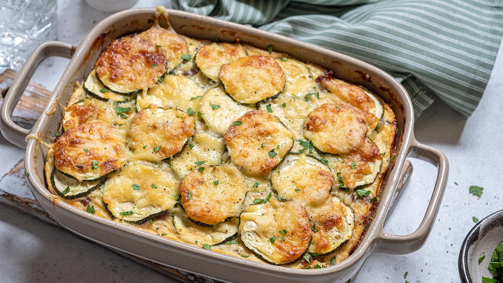 Auf dem Bild ist eine grüne Auflaufform zu sehen, randvoll gefüllt mit einem köstlichen Zucchini-Kartoffel-Auflauf. Die Oberfläche des Auflaufs ist goldbraun gebacken und sieht köstlich aus. Neben der Auflaufform liegt ein grün-weiß gestreiftes Tuch, das der Szene eine farbenfrohe Note verleiht. 