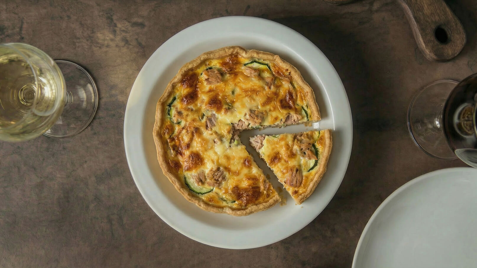 Eine gefüllte Quiche auf einem weißen Teller, mit goldbrauner, knuspriger Kruste und Stückchen von Zucchini und Fleisch. Ein Stück ist bereits herausgeschnitten. Der Teller steht auf einer Holztischoberfläche, umgeben von Glaswaren.