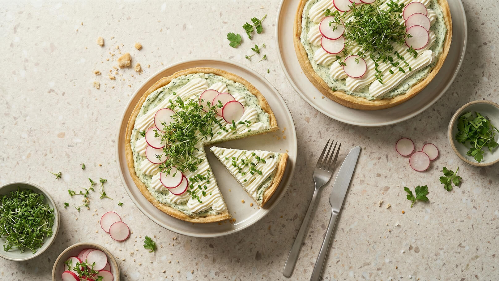 Ein runder, angesehener Kuchen, gefüllt mit einer cremigen Mischung, ist mit Radieschenscheiben, Kräutern und frischer Sahne verziert. Ein Stück wurde herausgenommen und auf einem Teller serviert. Neben dem Kuchen sind kleine Schalen mit frischen Kräutern und Radieschen.