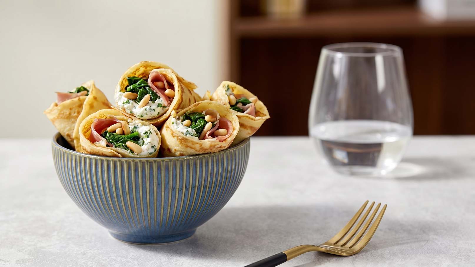 Eine Schüssel mit gefüllten Wraps liegt auf einem Tisch. Die Wraps sind mit Spinat, Schinken, und Käse gefüllt. Daneben steht ein Glas Wasser und eine Gabel. Der Hintergrund ist unscharf und zeigt einen modernen Wohnraum.