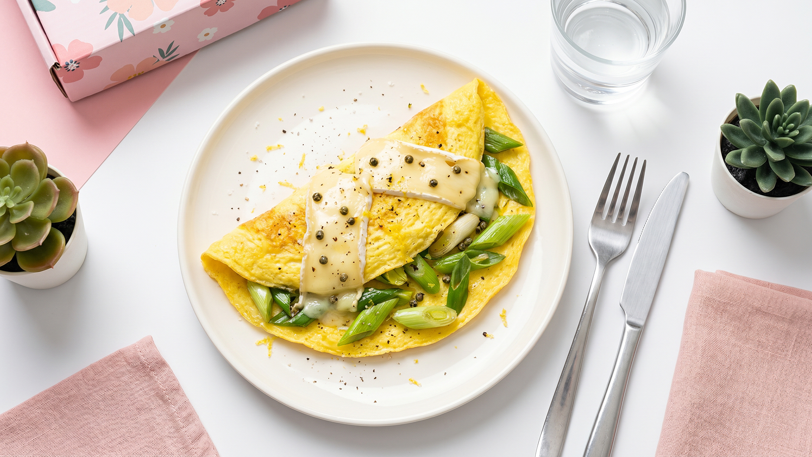Auf dem Bild ist ein Teller mit einer Omelette zu sehen, die mit Frühlingszwiebeln gefüllt ist und mit einer cremigen Sauce überzogen ist. Die Omelette ist gelb und auf einem weißen Teller angerichtet, umgeben von kleinen Pflanzen und einem Glas Wasser.
