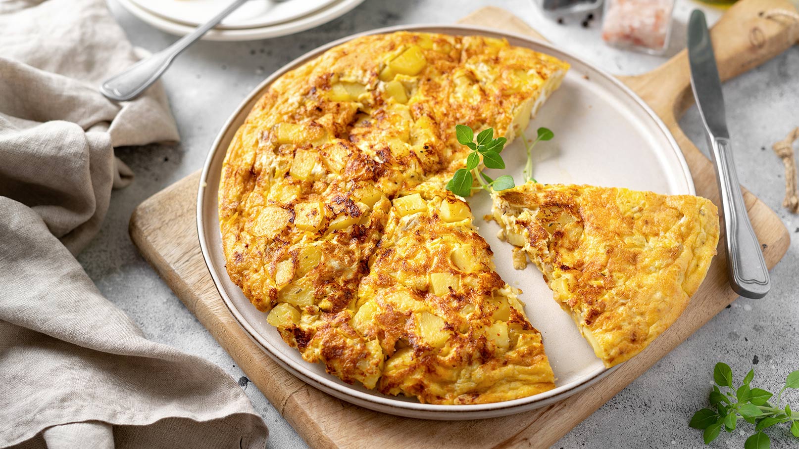 Auf dem Bild ist ein großer weißer Teller zu sehen, auf dem ein saftiges Kartoffel-Omelett mit Käse serviert wird. Das Omelett ist goldbraun gebraten und mit geschmolzenem Käse überbacken. Der Teller steht auf einem rustikalen Holzbrett, auf dem ein Messer liegt. Die natürliche Holzstruktur des Brettes und das klare Weiß des Tellers bilden einen schönen Kontrast und verleihen dem Gericht eine ansprechende Präsentation.