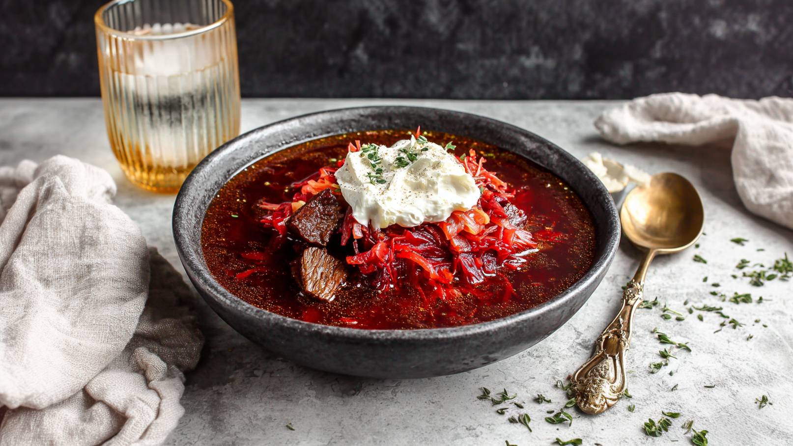 Eine Schüssel mit Borschtsch, gefüllt mit rotem Kraut und Stückchen von Fleisch, garniert mit einem Klecks saurer Sahne und frischen Kräutern. Neben der Schüssel liegt ein goldfarbener Löffel auf einer grauen Unterlage. Im Hintergrund steht ein Glas mit Wasser.