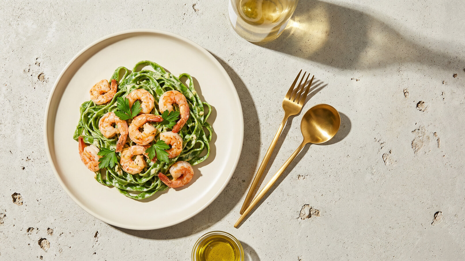 Ein Teller mit grüner Pasta, garniert mit gebratenen Garnelen und frischer Petersilie. Im Hintergrund befinden sich eine Gabel, ein Löffel und ein Glas Weißwein.