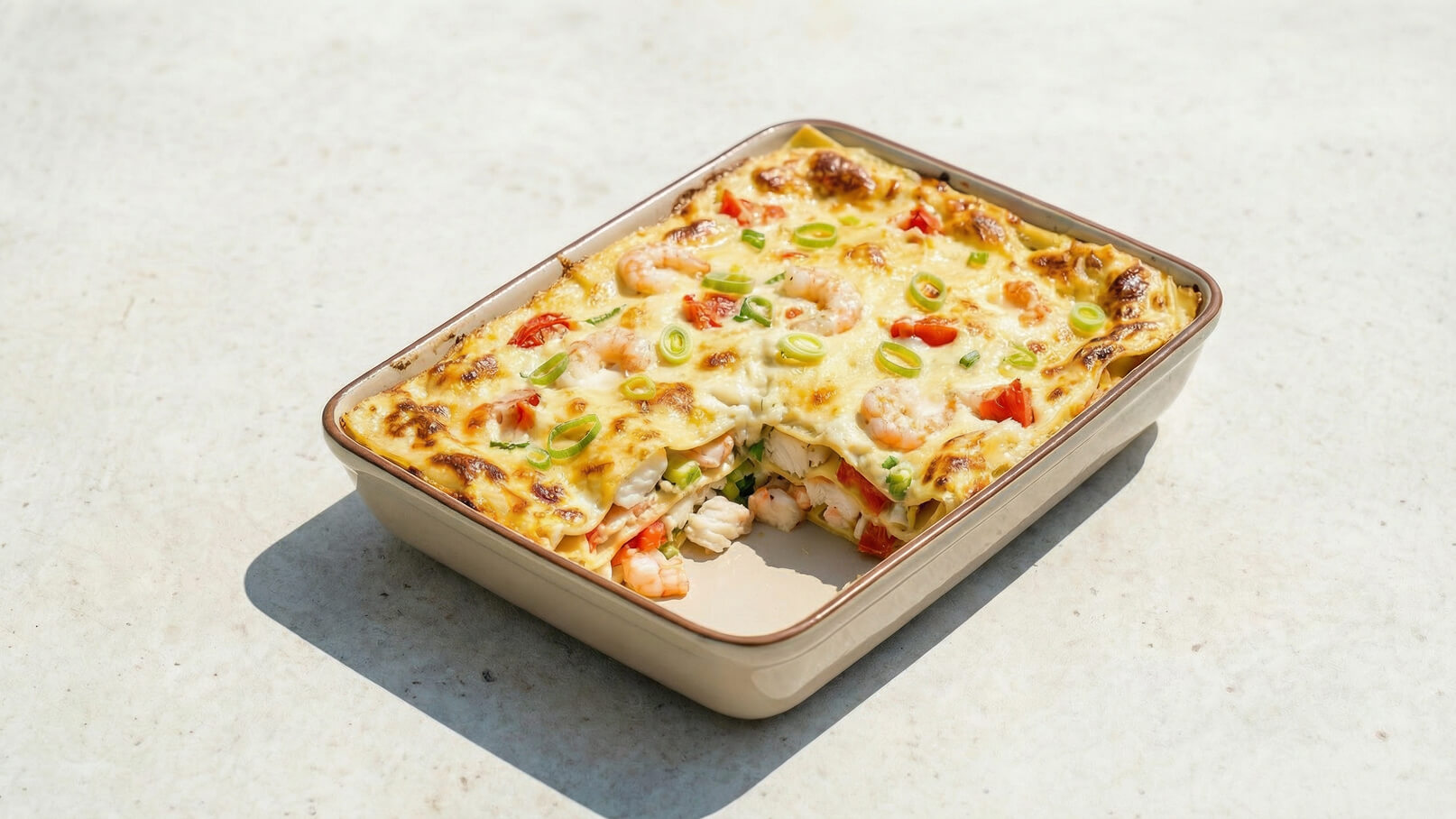 Eine Auflaufform mit Lasagne, die mit Garnelen, Tomaten und Frühlingszwiebeln belegt ist. Die oberste Schicht ist goldbraun und leicht knusprig. Ein Stück Lasagne ist herausgenommen, sodass die Schichten sichtbar sind.