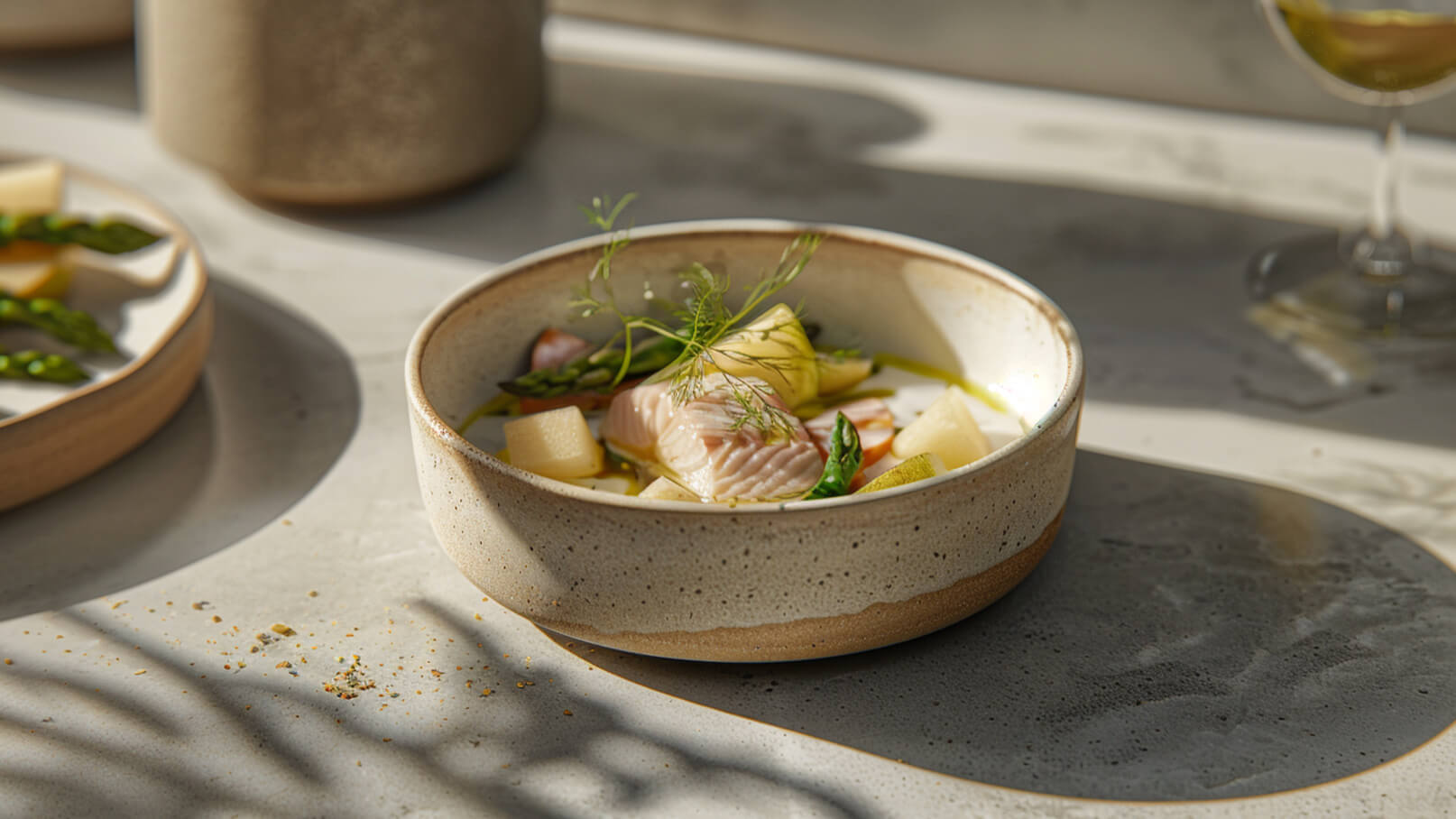 Ein helles Keramikschälchen enthält ein Gericht mit Fischstücken, Spargel und verschiedenen Gemüsestücken, angereichert mit einem Essig- oder Öl-Dressing. Der Teller steht auf einer glatt strukturierten Oberfläche, die von sanftem Licht beleuchtet wird.