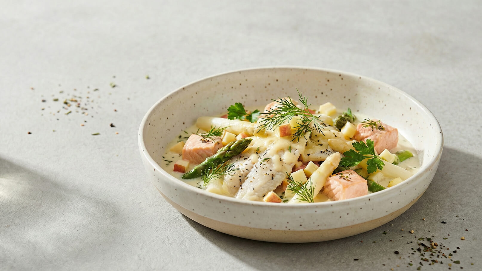 Eine Schüssel mit cremigem Gericht, das Stücke von Lachs, Spargel und Kartoffeln enthält, garniert mit frischem Dill und Petersilie. Der Hintergrund ist hell und schlicht, Ablagerungen von Kräutern sind sichtbar, aber nicht dominant.