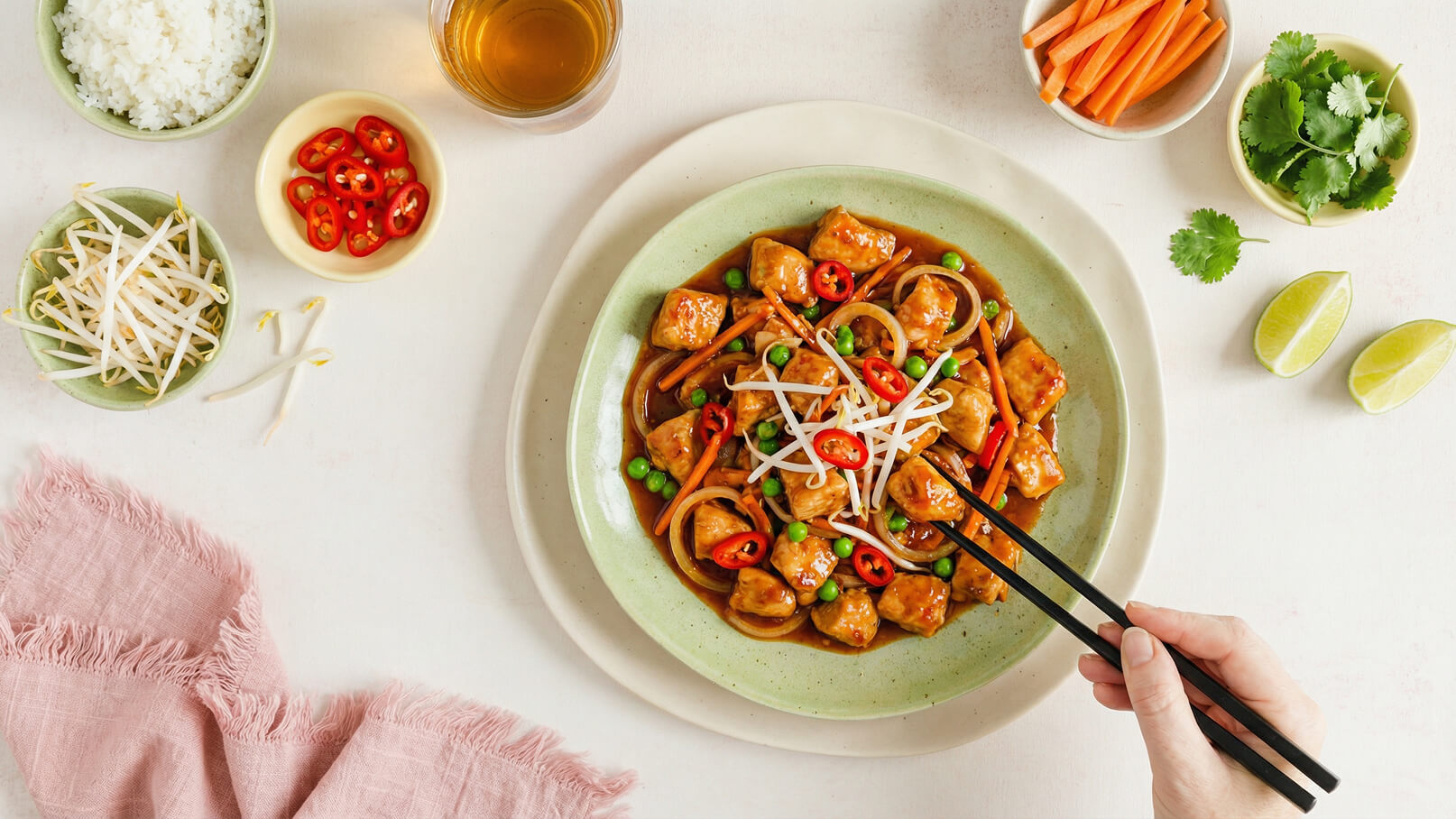 Ein Gericht mit gebratenem Tofu und buntem Gemüse, garniert mit Sojasprossen und roten Chili-Scheiben auf einem grünen Teller. Daneben befinden sich Schalen mit Reis, frischen Karotten, Limettenscheiben und Koriander. Eine Hand greift mit Essstäbchen nach dem Essen.
