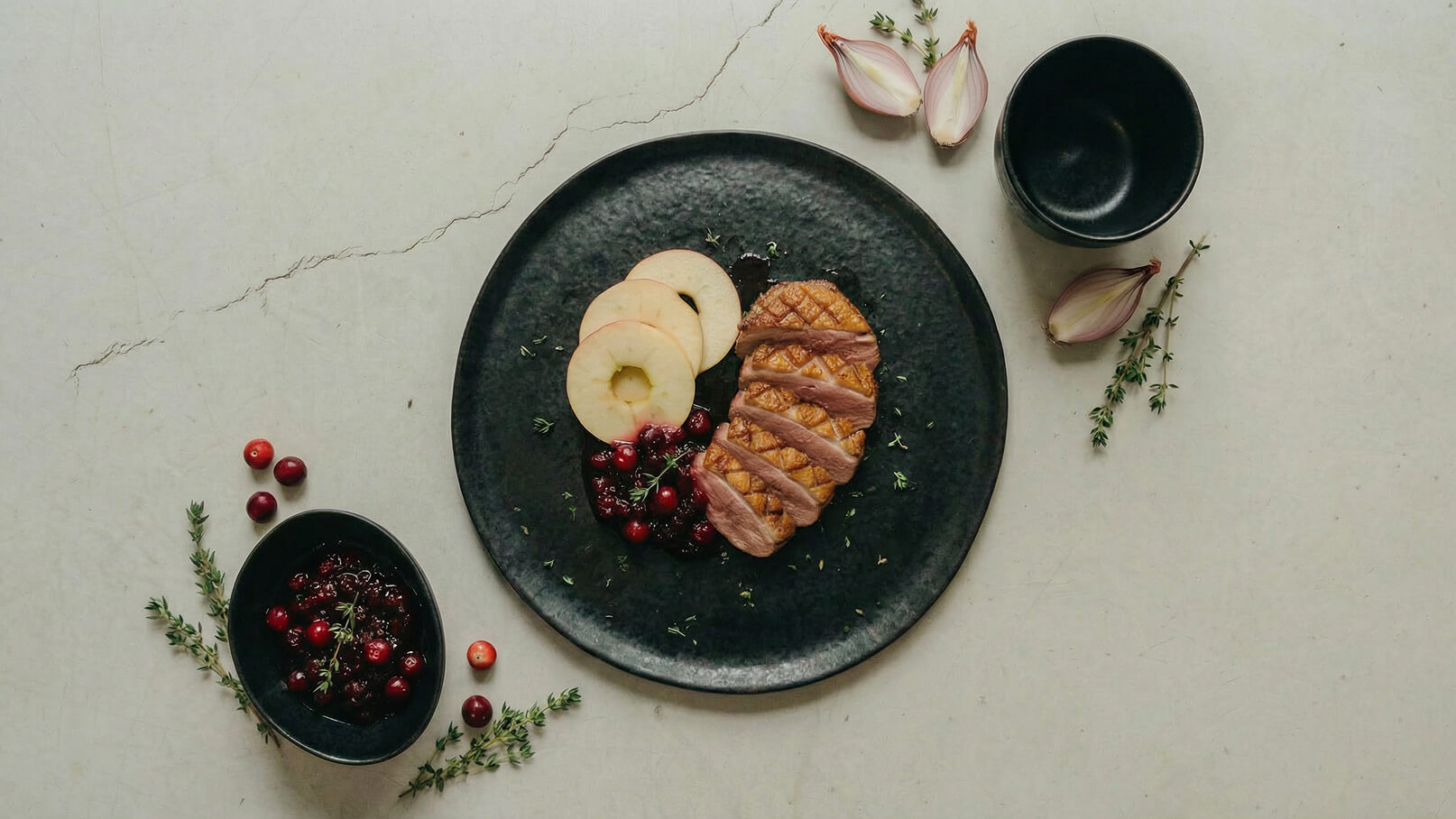 Auf einem schwarzen Teller befindet sich gegrilltes Fleisch, garniert mit frischen Thymianzweigen und umgeben von Apfelscheiben und einer Cranberry-Sauce. Neben dem Teller liegen einige ganze Cranberries und Schalotten.