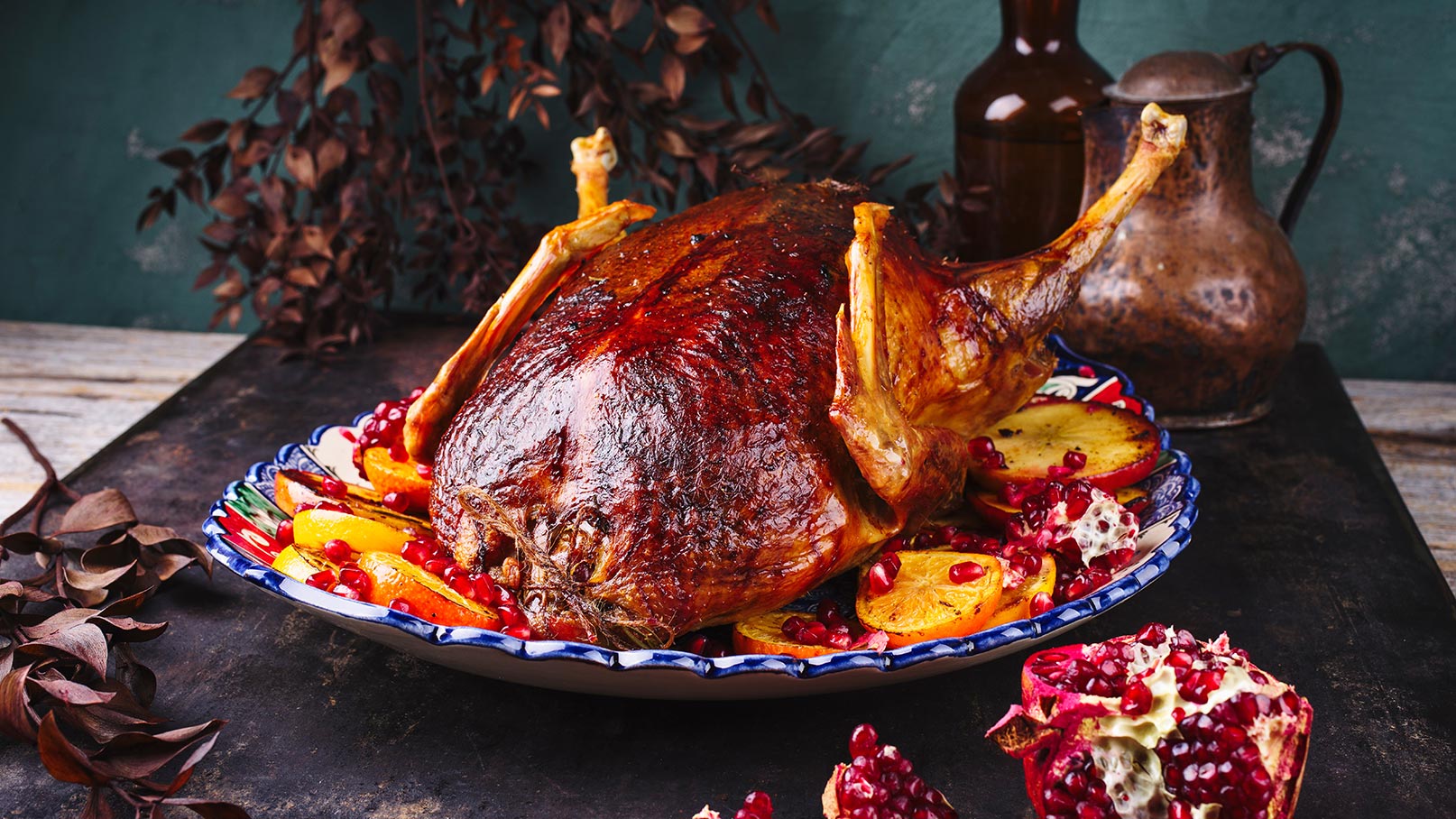 Das Bild zeigt einen eleganten blau-weißen Teller mit einem saftigen Gänsebraten, der eine goldene, knusprige Haut hat. Eine Orangenscheibe fügt einen Farbtupfer hinzu. Im Vordergrund liegt ein halber Granatapfel, dessen tiefrote Kerne einen schönen Kontrast bilden. Die Gesamtkomposition wirkt festlich und appetitlich.