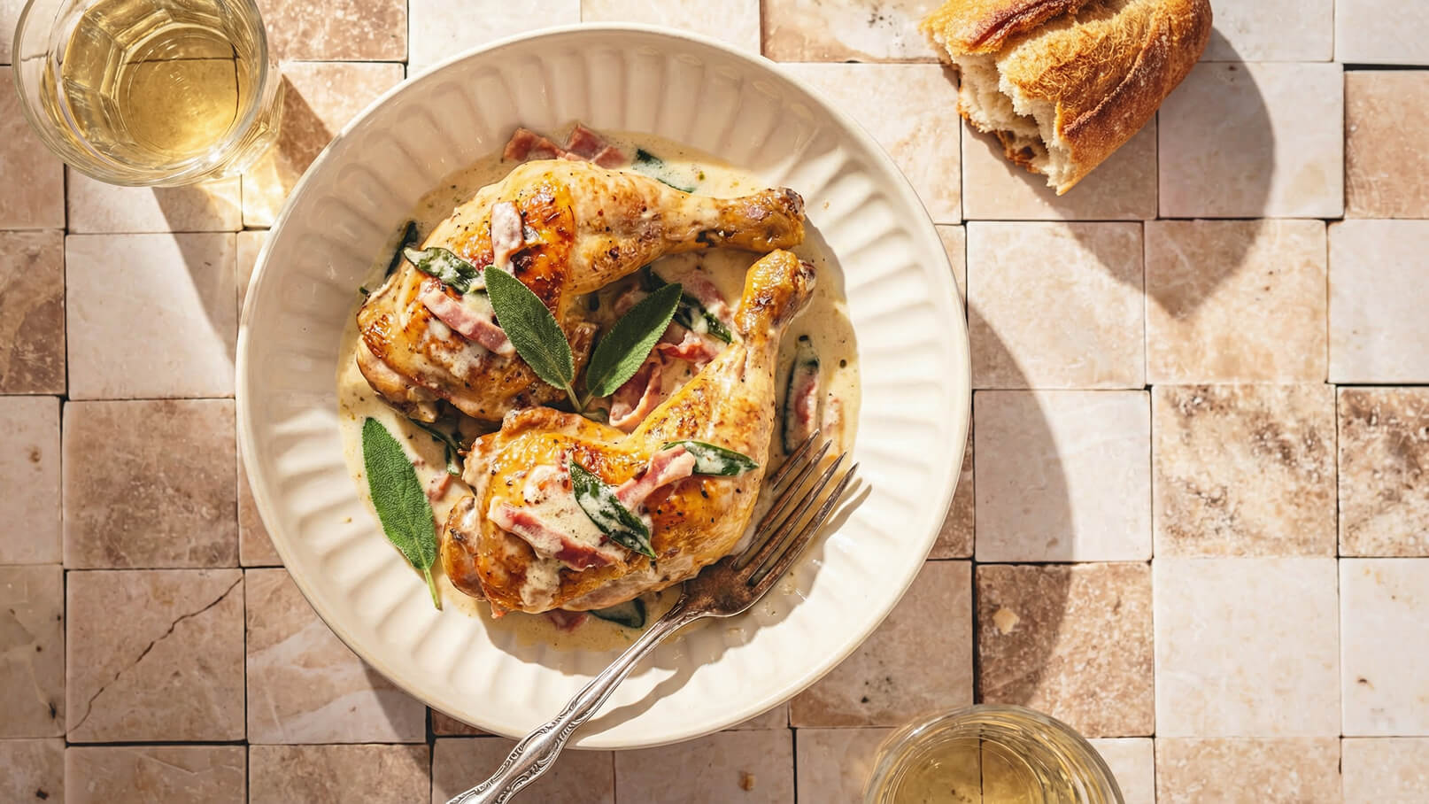 Ein tiefes Teller mit zwei saftigen Hähnchenkeulen in einer cremigen Soße, garniert mit Schinken und Salbei. Dazu liegt ein Stück abgebrochenes Brot auf einem Tisch aus hellen Fliesen. Im Hintergrund steht ein Gläser mit hellem Getränk.