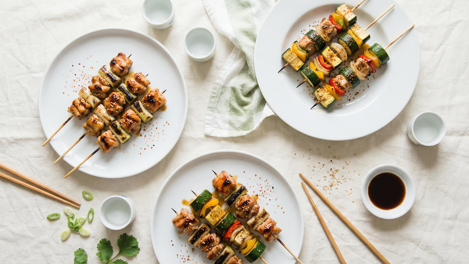 Drei Teller mit Grillspießen sind auf einem hellen Tisch angeordnet. Jeder Spieß enthält mariniertes Fleisch und Gemüse wie Zucchini, Paprika und Zwiebeln. Dazu gehören Schalen mit Sojasauce und kleinen Gläsern. Grüne Lauchzwiebeln und frische Kräuter sind ebenfalls sichtbar.