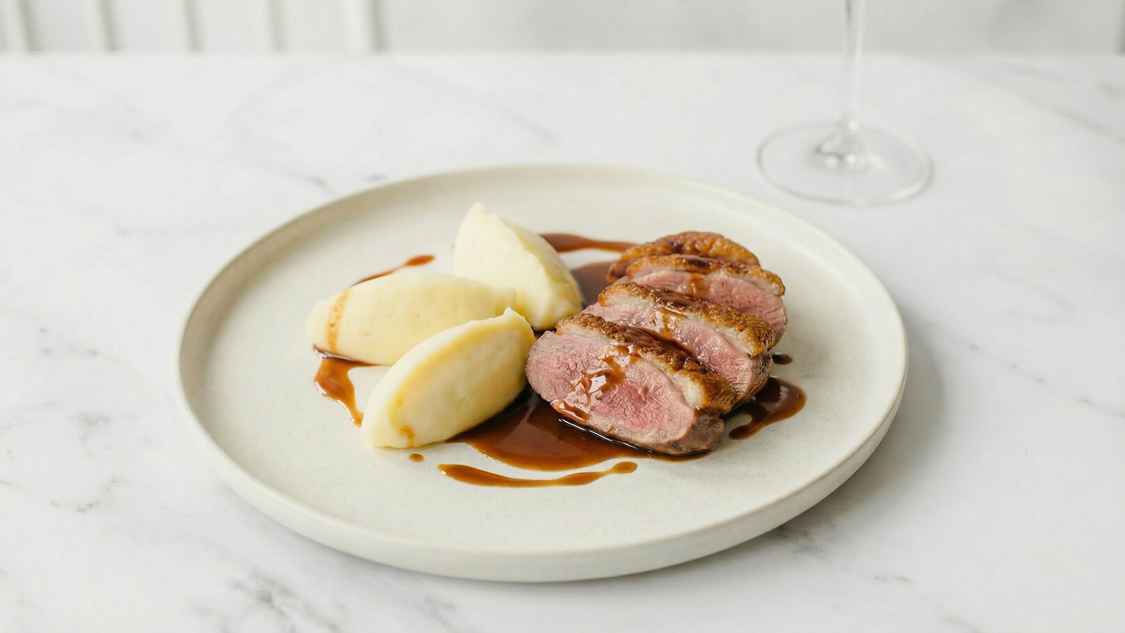 Ein Teller mit dünn geschnittenem, saftigem Entenbrustfilet, garniert mit einer dunklen Sauce. Daneben liegen zwei Portionen cremiger Kartoffelpüree. Der Teller hat einen schlichten, hellen Rand und die Hintergrundfläche ist neutral gehalten.