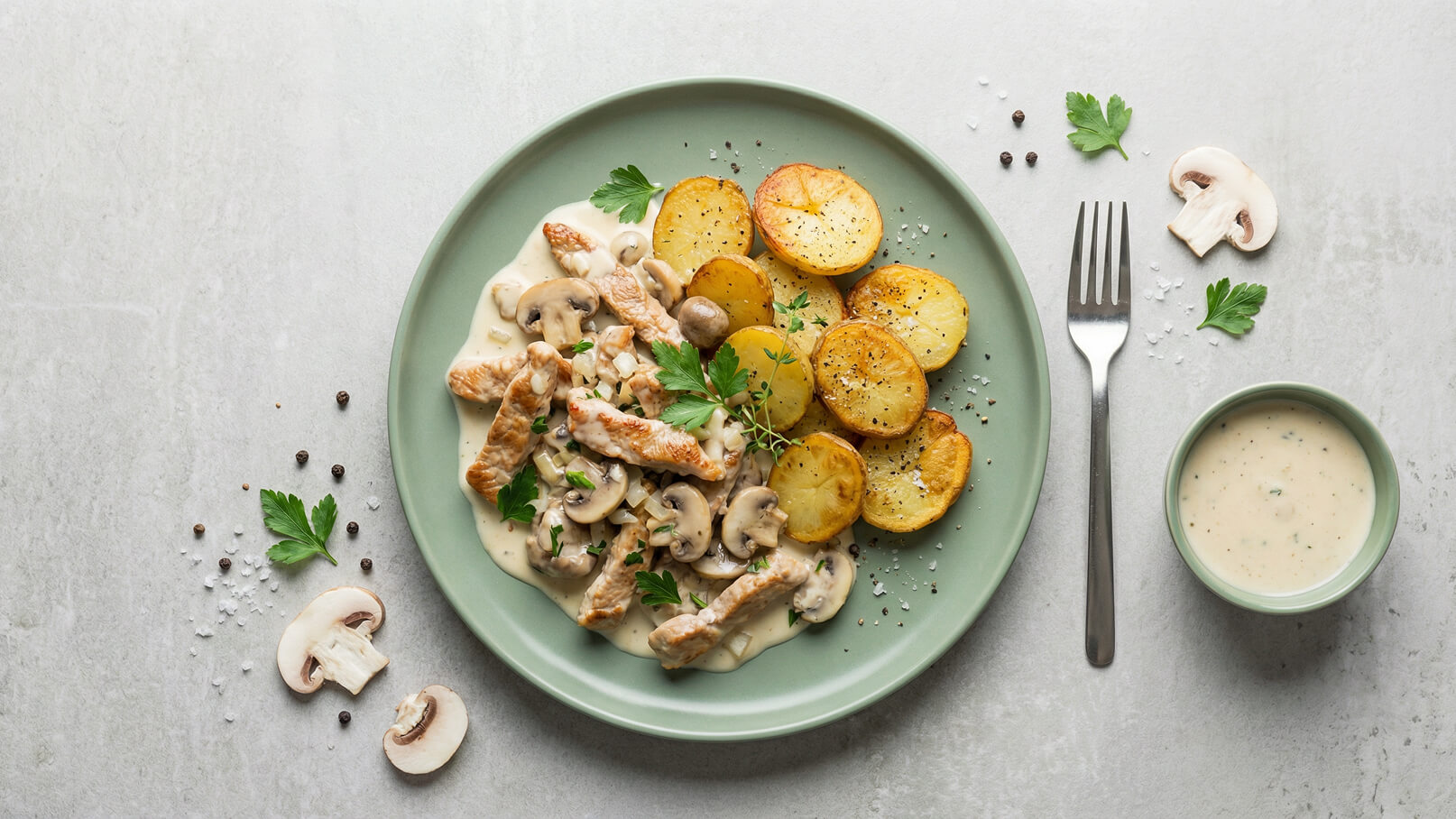 Ein Teller mit zarten Hähnchenstreifen in einer cremigen Sauce, garniert mit frischen Kräutern und Pilzen, daneben knusprige, goldbraun gebratene Kartoffelscheiben. Ein kleines Schälchen mit heller Sauce ist ebenfalls vorhanden.