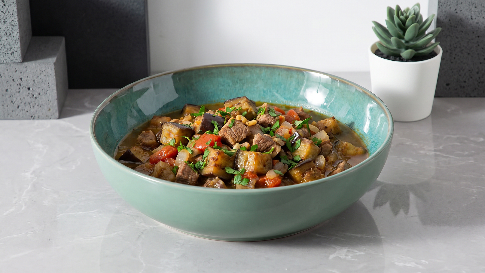 Eine große Schüssel mit einer bunten Mischung aus Gemüse und Fleisch, darunter Zucchini, Tomaten und Kartoffeln, die in einer würzigen Sauce serviert werden. Die Schüssel hat einen türkisfarbenen Rand und steht auf einer marmorierten Oberfläche.