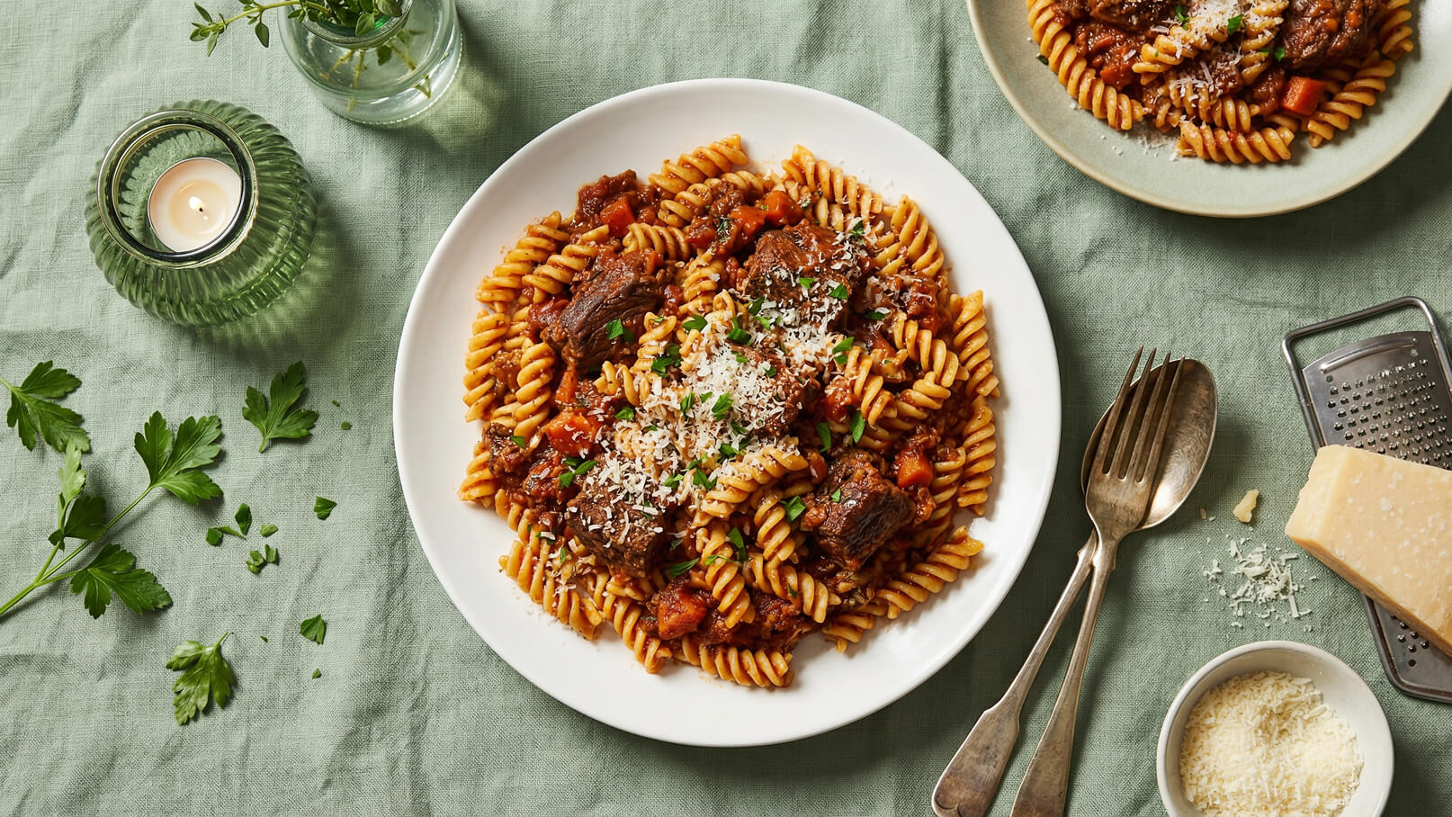 Ein Teller gedrehte Pasta mit Rinderfleisch und Tomatensauce, garniert mit frischem Parmesan und Kräutern. Daneben liegen eine Gabel, ein Löffel und eine Handvoll geriebener Parmesan. Der Hintergrund ist in sanften Grüntönen gehalten.