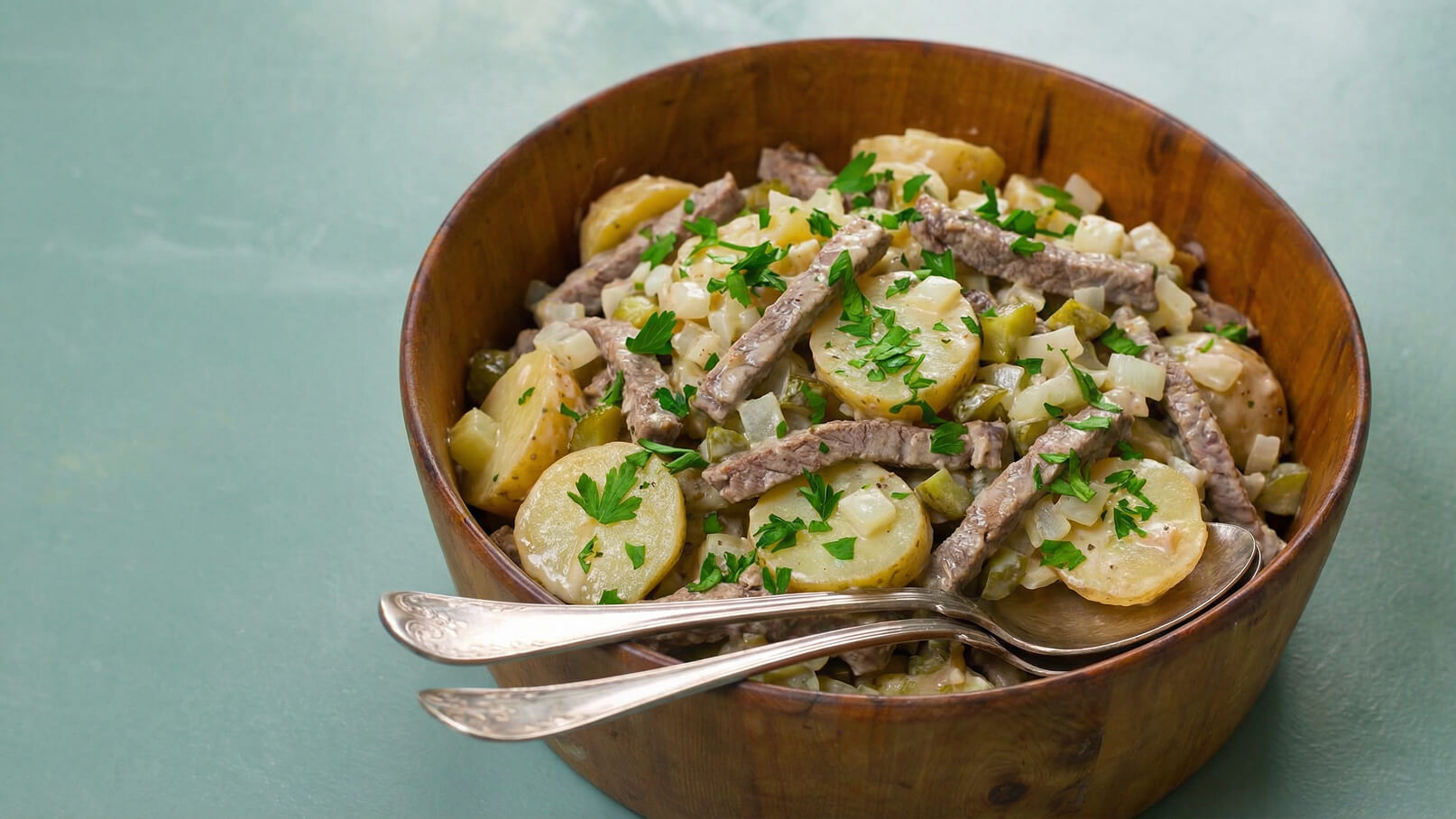 Eine Holzküche voller Salat mit Kartoffeln, Fleischstreifen und gehackter Petersilie. Zwei Silberlöffel liegen neben der Schüssel. Der Salat erscheint frisch und appetitlich.