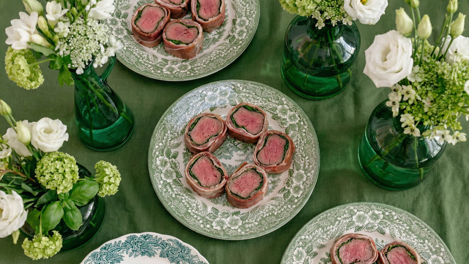 Auf dem Tisch liegen mehrere Teller mit Schalen, die gefüllte Fleischröllchen zeigen. Die Röllchen bestehen aus magerem Rindfleisch, umgeben von grünen Blättern. Der Tisch ist mit einer grünen Tischdecke dekoriert und mit kleinen Blumenvasen geschmückt.