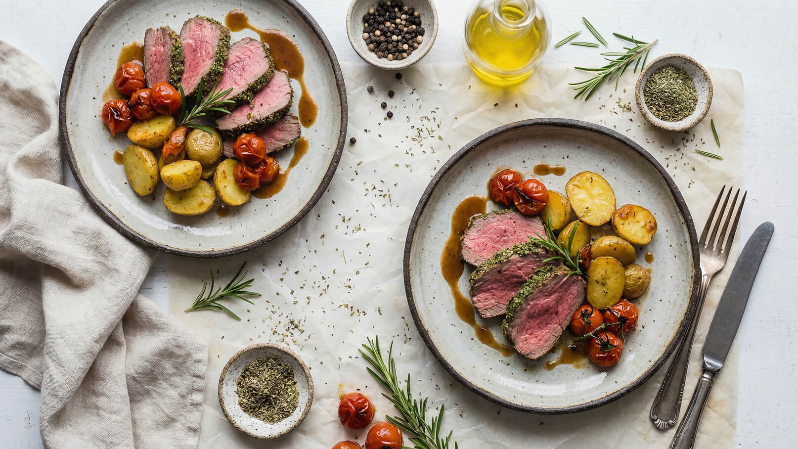 Zwei elegante Teller mit gebratenem Rindfleisch, serviert mit goldenen Kartoffeln und gerösteten Tomaten. Frischer Rosmarin verleiht das Aroma. Daneben liegen ein kleines Gefäß mit Gewürzen und ein Glas mit Olivenöl. Der Tisch ist mit einem hellen Tuch und dekorativen Kräutern angerichtet.