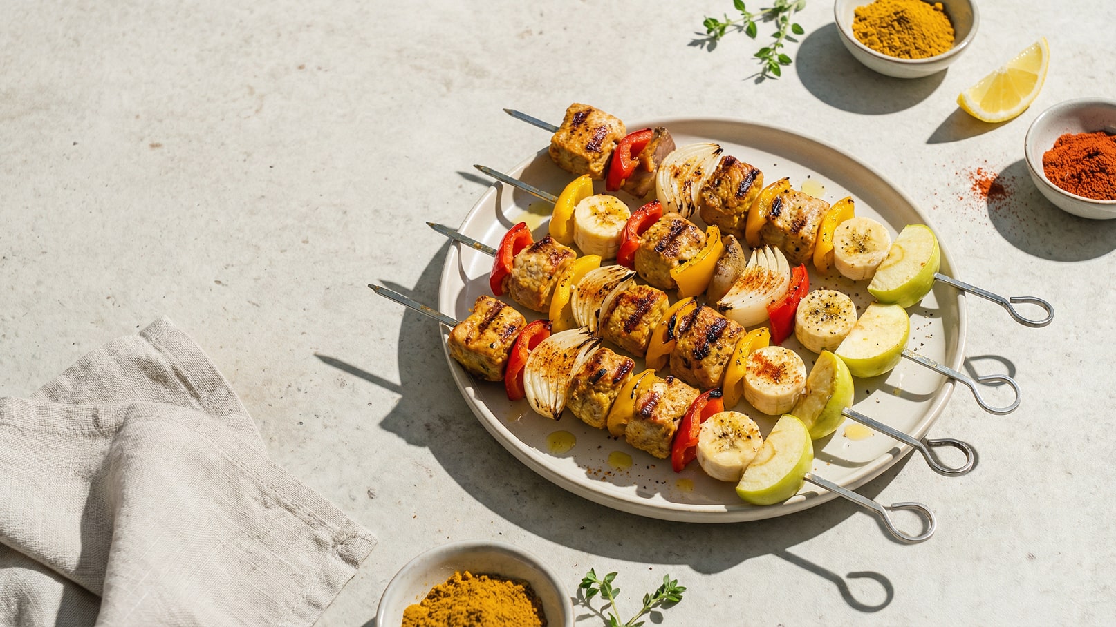 Ein Teller mit bunten Grillspießen, bestehend aus Fleisch, Paprika, Zwiebeln, Bananen und Äpfeln. Die Spieße sind appetitlich angerichtet und leicht angegrillt. Neben dem Teller liegen Gewürze und eine Zitronenscheibe.
