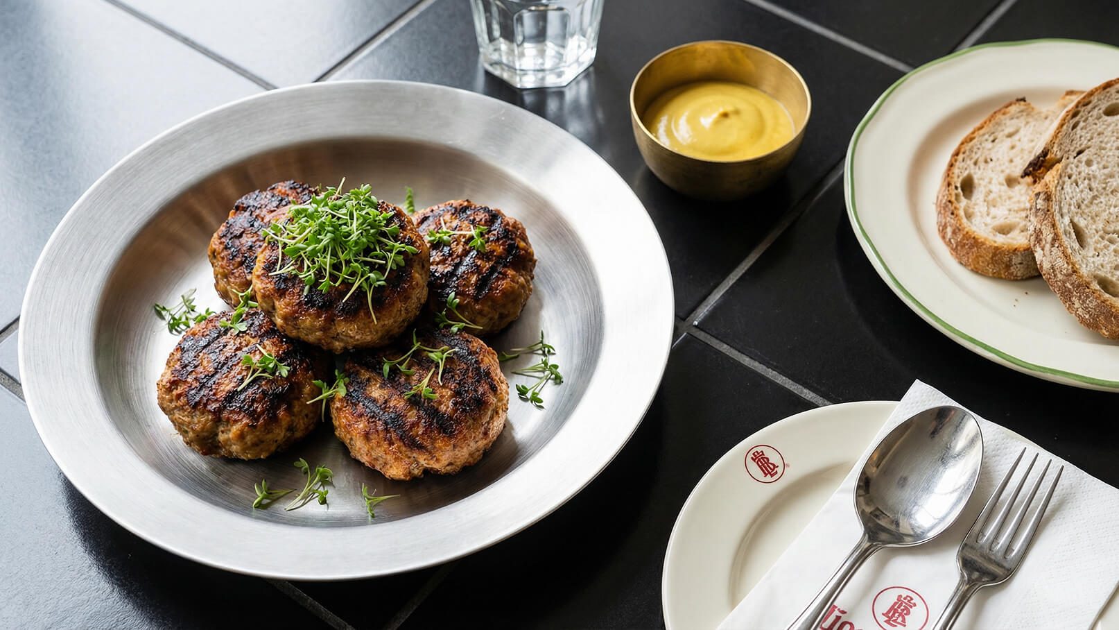 Auf einem silbernen Teller liegen drei gegrillte Frikadellen, garniert mit frischen Kräutern. Daneben befindet sich eine Schale mit Senf und ein kleiner Teller mit zwei Scheiben Brot. Der Hintergrund ist dunkel und schlicht gehalten.