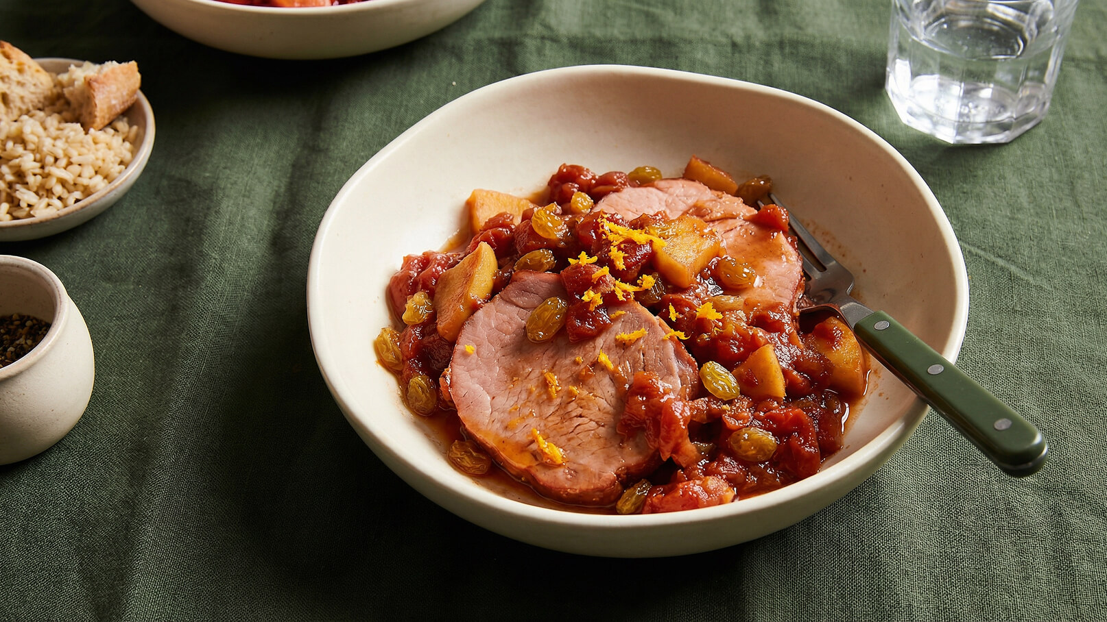 Ein tiefes Teller mit einer Portion Fleisch, umgeben von einer fruchtigen Sauce aus Tomaten, Äpfeln und Rosinen. Die Speise ist mit orangefarbener Schale garniert. Im Hintergrund sind weitere Schüsseln mit Beilagen und ein Glas Wasser sichtbar, auf einem grünen Tisch.