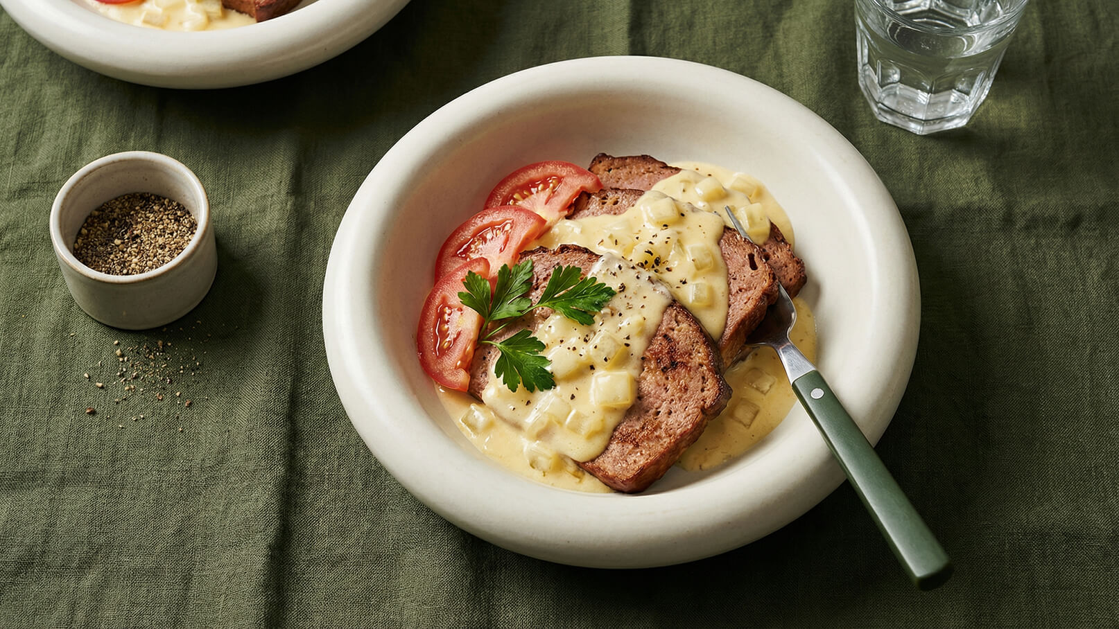 Ein Teller mit angerichtetem Fleisch, überzogen mit einer cremigen Sauce, und garnierter mit frischen Tomatenscheiben und Petersilie. Daneben steht eine kleine Schüssel mit Pfeffer sowie ein Glas Wasser. Der Hintergrund ist grün und vermittelt eine einladende Atmosphäre.