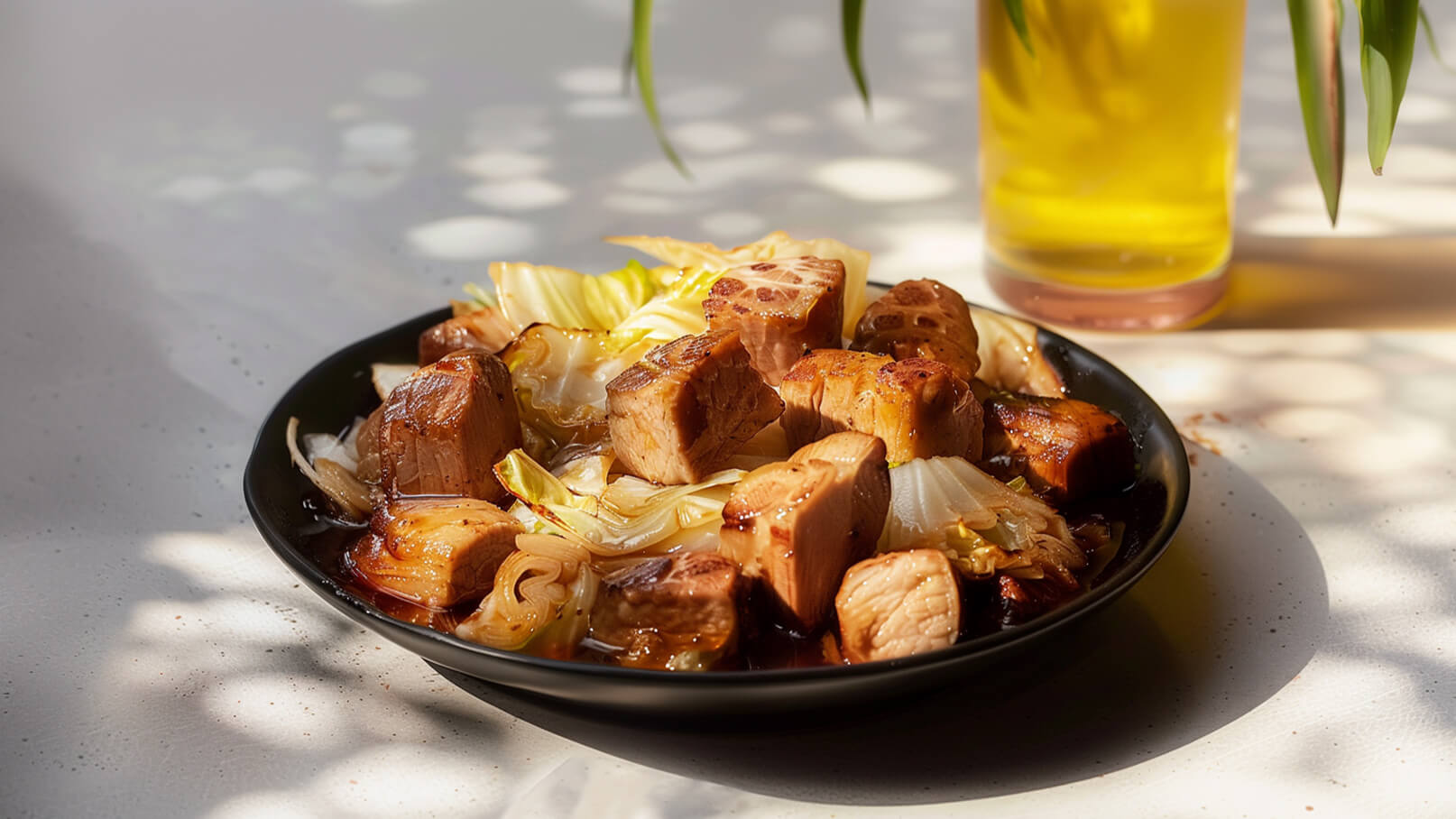 Ein schwarzer Teller mit zarten Stückchen Fleisch und gebratenem Gemüse, darunter frischer Kohl und Zwiebeln, in einer schmackhaften Sauce. Hinter dem Teller steht ein Glas mit einem goldenen Getränk, das dem Gericht eine einladende Atmosphäre verleiht.