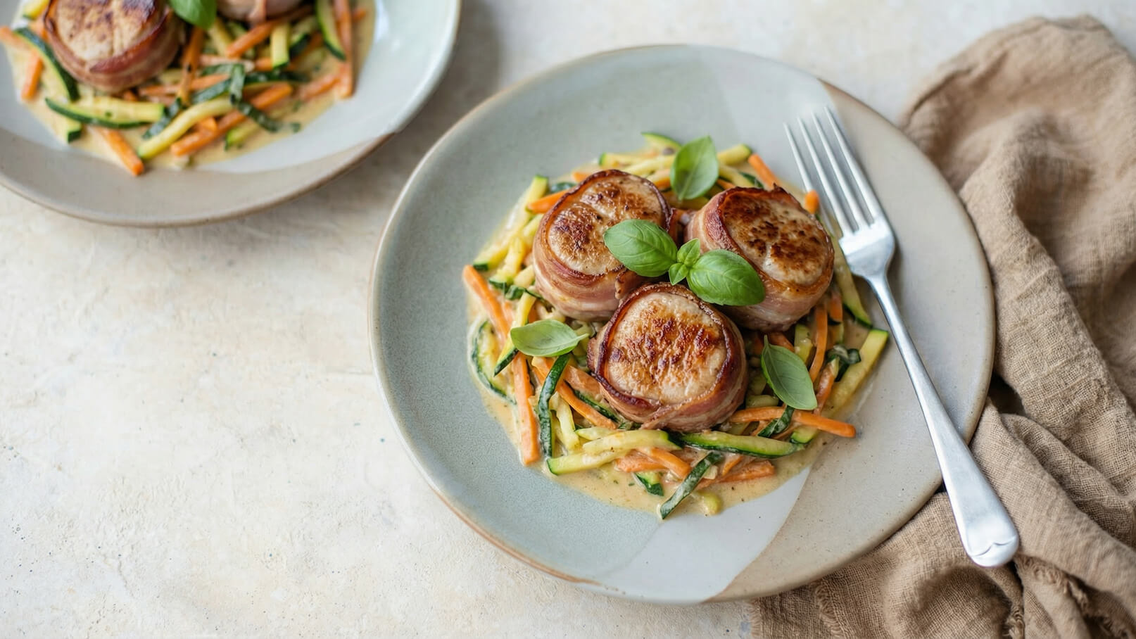 Auf dem Teller liegen drei in Speck umwickelte Medaillons auf einem Bett aus Gemüse, darunter Zucchini und Karottenstreifen. Das Gericht ist leicht mit einer cremigen Sauce überzogen und mit frischen Basilikumblättern garniert. Ein weiteres ähnliches Gericht liegt im Hintergrund.