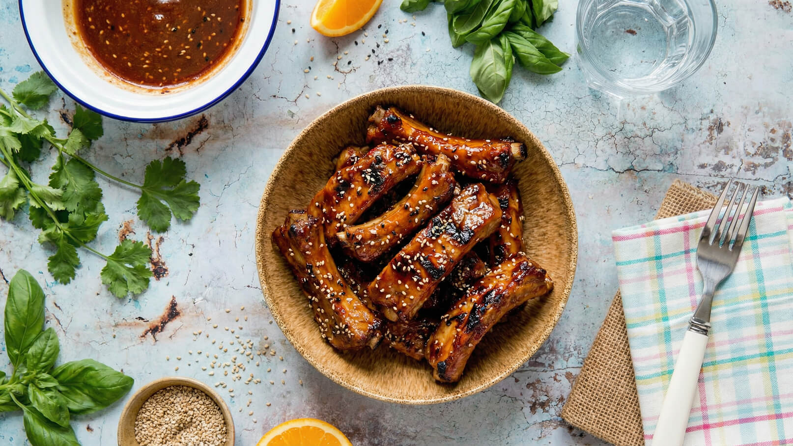 Eine Schüssel mit glasierten Rippchen, bestreut mit Sesam, steht auf einem rustikalen Tisch. Neben der Schüssel befinden sich frische Kräuter, ein Glas Wasser, ein Handtuch und eine Gabel. Im Hintergrund ist eine Schale mit Sauce und eine halbe Orange zu sehen.