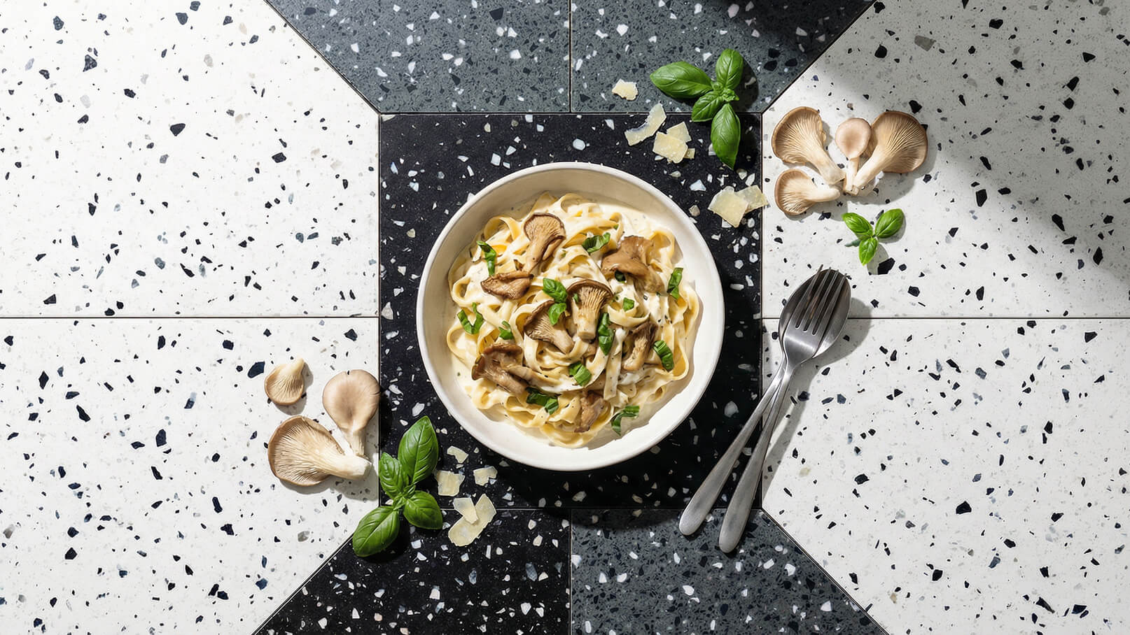 Ein Teller Pasta mit cremiger Sauce, belegt mit Pilzen und frischem Basilikum. Der Teller ist auf einer stylischen, gemusterten Oberfläche platziert. Neben dem Teller liegen einige frische Pilze und Basilikumblätter. Ein Besteckset ist ebenfalls zu sehen.