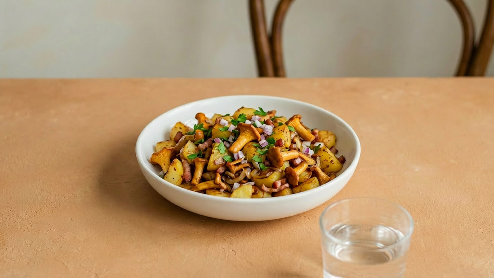 Eine weiße Schüssel mit einer Mischung aus gebratenen Kartoffeln, Pfifferlingen und kleinen Stückchen Speck. Die Speise ist mit frischer Petersilie und kleinen Zwiebelwürfeln garniert. Im Hintergrund steht ein Glas Wasser auf einem beige-farbenen Tisch.