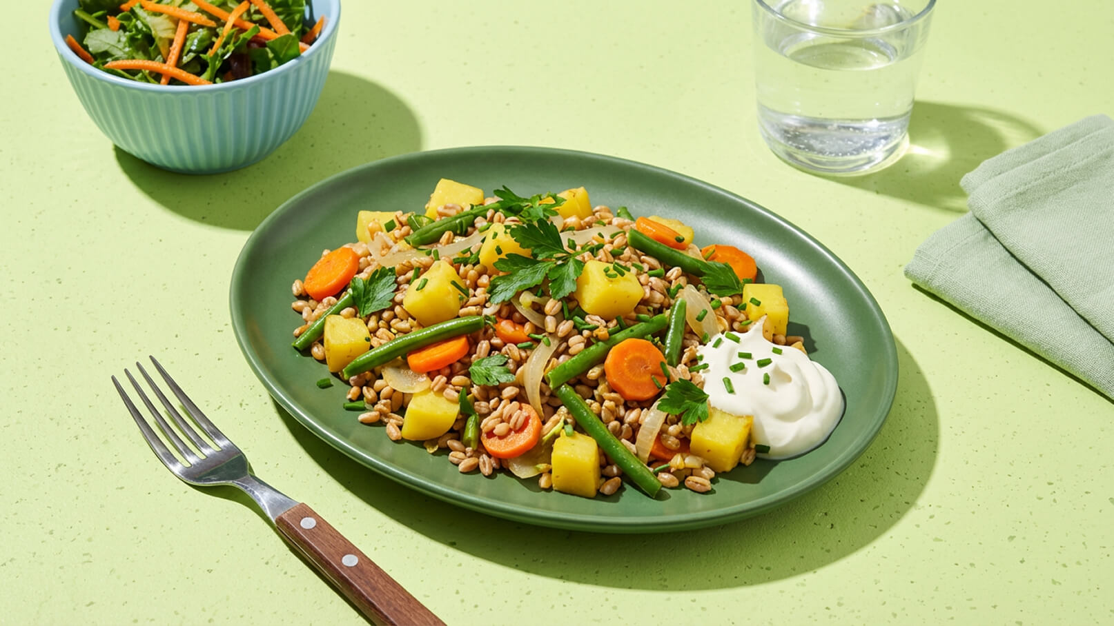 Ein grüner Teller mit einer Mischung aus Dinkel, Kartoffeln, Karotten und grünen Bohnen, garniert mit Kräutern und einem Klecks Joghurt. Daneben ein Schalen Salat mit Karottenstreifen und ein Glas Wasser. Der Hintergrund ist in einem sanften Grünton.