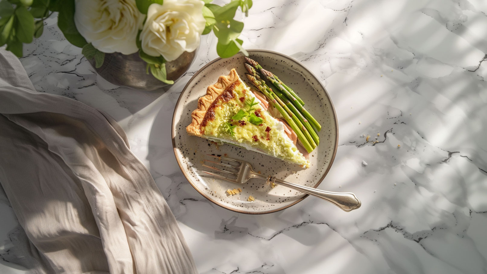 Ein Teller mit einem Stück Quiche, garniert mit frischen Kräutern, und einer Beilage von grünem Spargel. Der Hintergrund zeigt eine elegante, marmorierte Oberfläche. Ein Besteck liegt neben dem Teller.