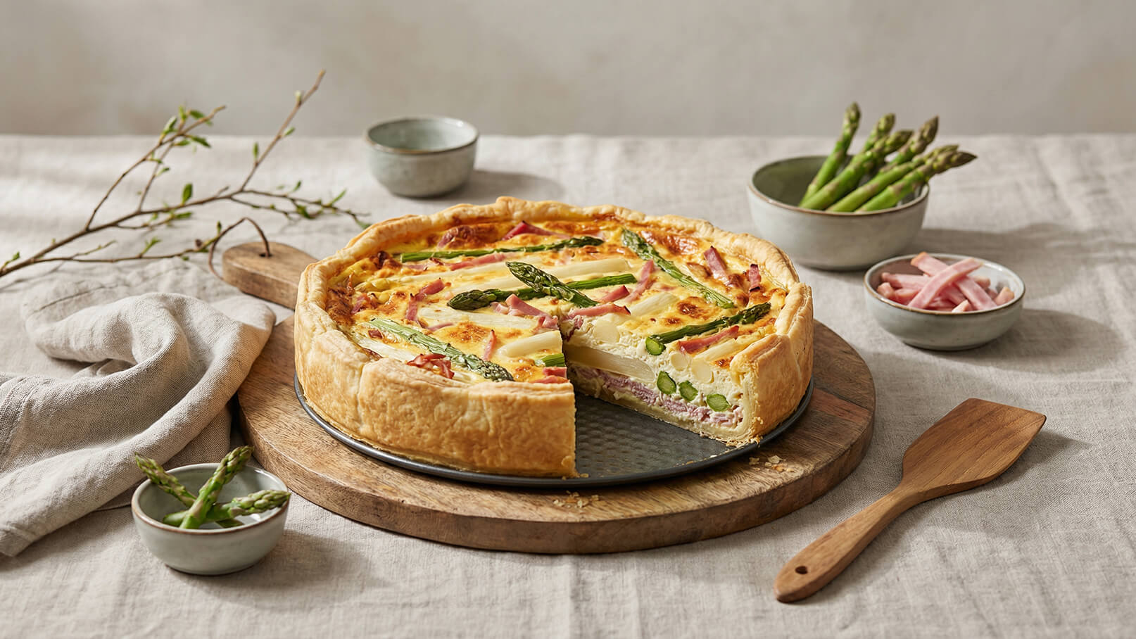 Ein frisch gebackener Quiche mit Spargel und Schinken auf einem Holzbrett. Ein Teil der Quiche ist herausgeschnitten, und daneben stehen kleine Schalen mit weiteren Spargelstücken und Schinken. Im Hintergrund sind Tabletts und ein Textil sichtbar.
