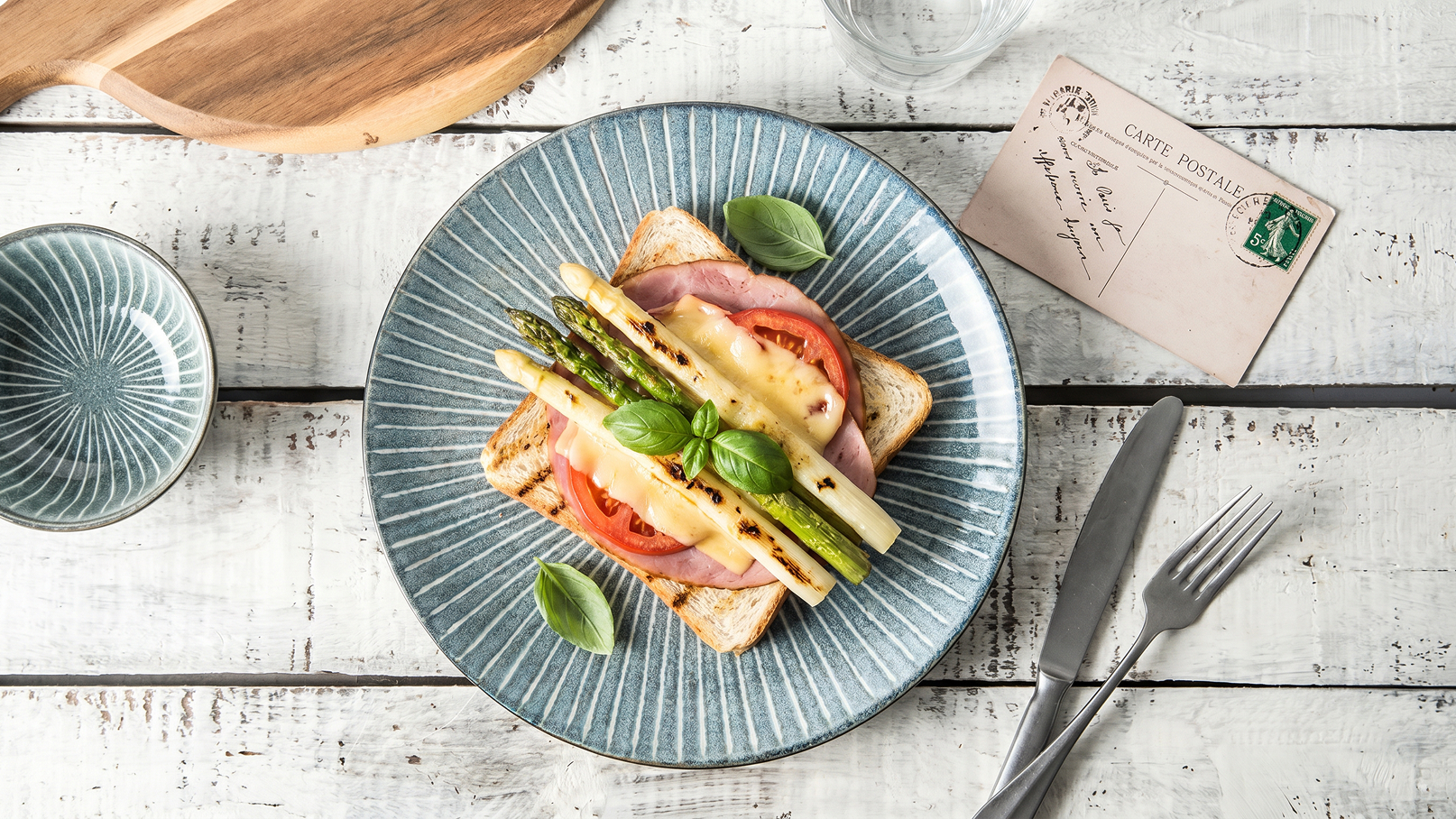 Ein Teller mit geröstetem Brot, belegt mit Schinken, Tomaten und gegrilltem Spargel. Darauf liegt geschmolzener Käse und frische Basilikumblätter. Neben dem Teller steht eine kleine Schüssel und eine Postkarte. Der Hintergrund ist aus hellem Holz.