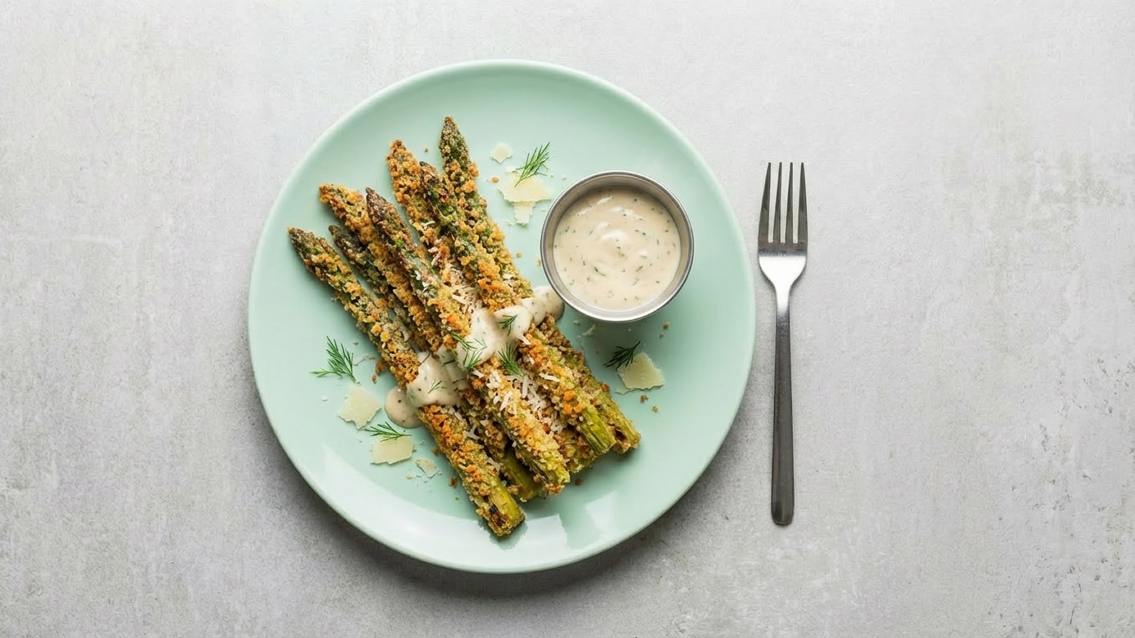 Auf einem mintgrünen Teller liegen mehrere Portionen panierter Spargel, garniert mit einer cremigen Sauce. Neben dem Teller befindet sich eine Gabel. Der Hintergrund ist hell und neutral gehalten.